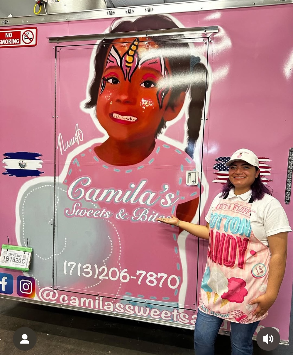 A woman in a white cap and apron stands next to a pink food truck with a cartoon face of a girl with a unicorn horn, smiling, pointing at the truck's sign. The truck displays the name 'Camila's Sweets & Bites' and contact information.