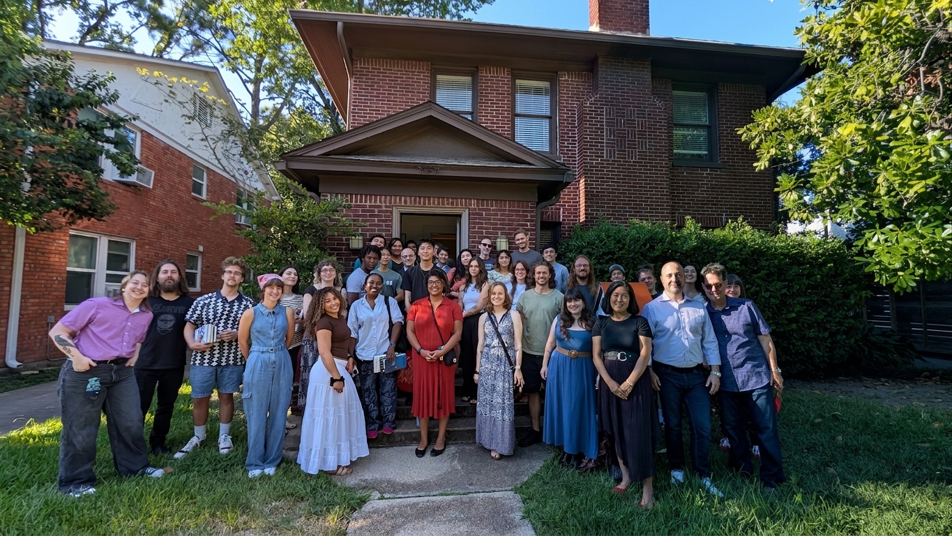 Group of people standing on a sidewalk in front of a brick house with trees and bushes, sunny day.