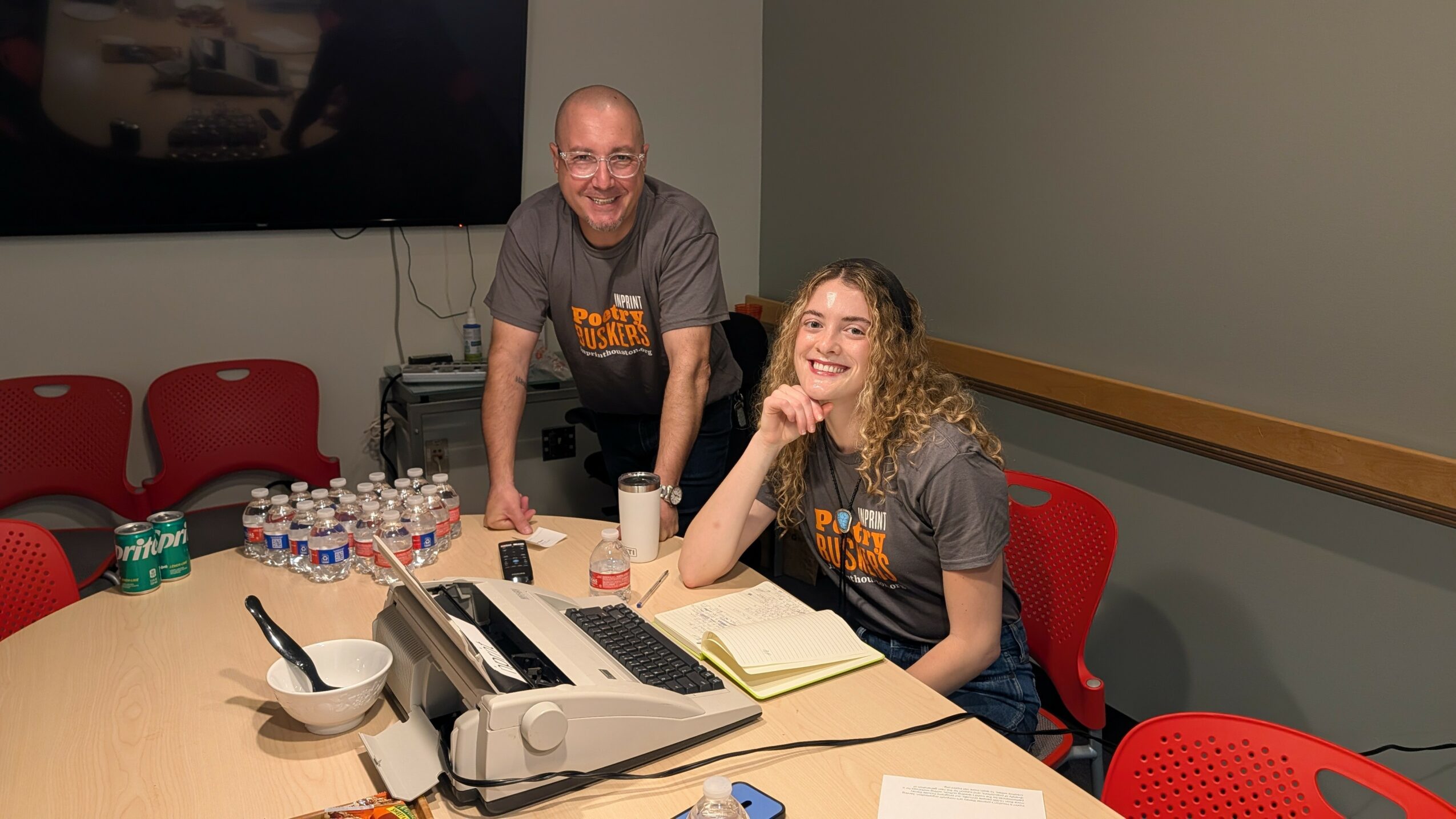 Two people in gray shirts sit at a conference table with phones, notebooks, and water bottles, smiling in a meeting room.