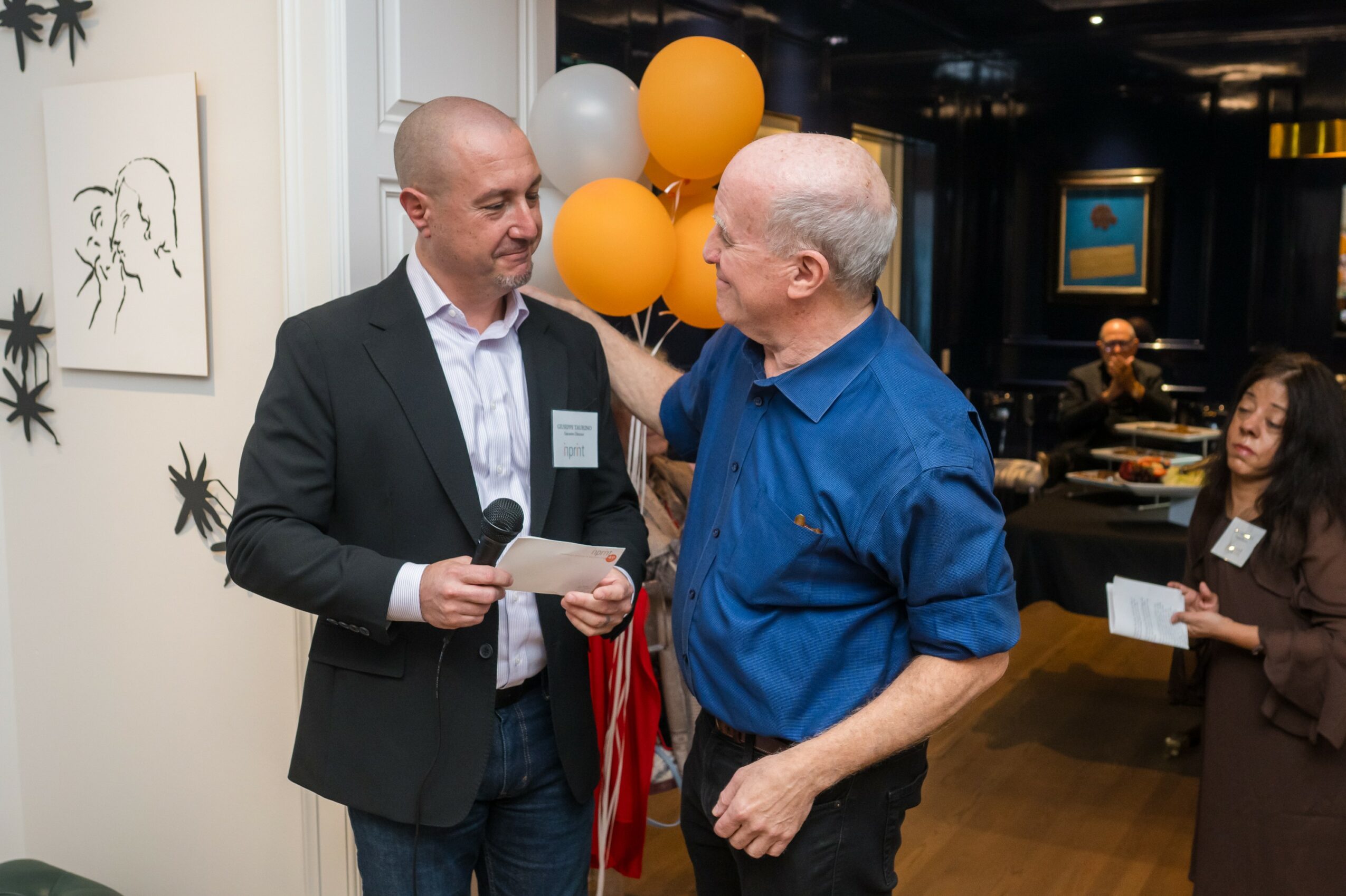 Two men talking at an indoor event, one holding a paper, with balloons and artwork in the background.