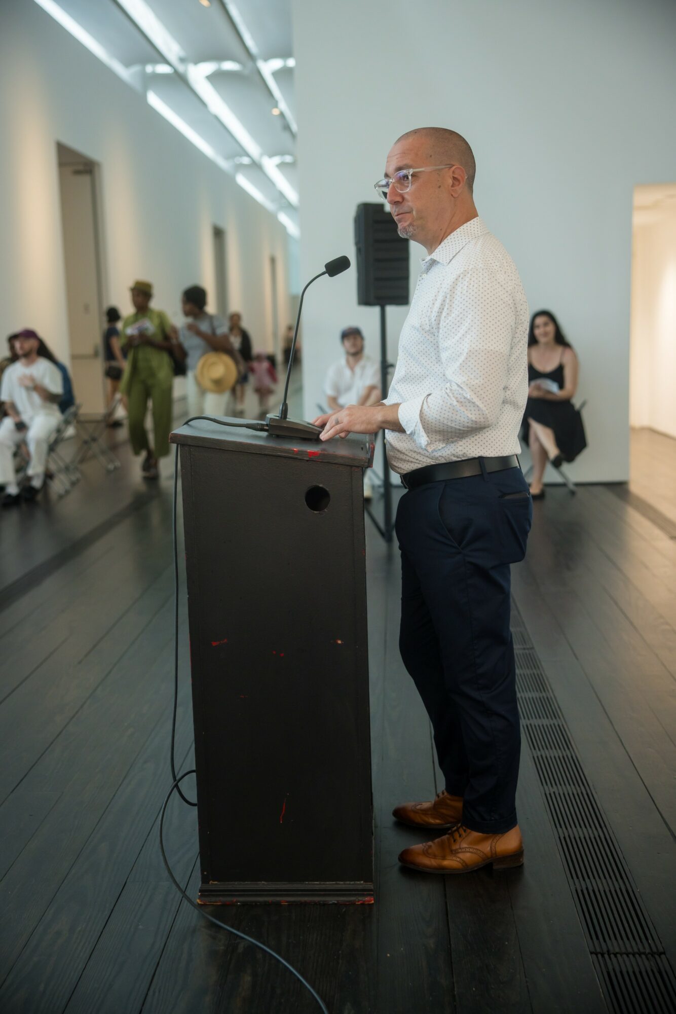 Man in white shirt and dark pants speaking at a podium in a gallery with people in the background.