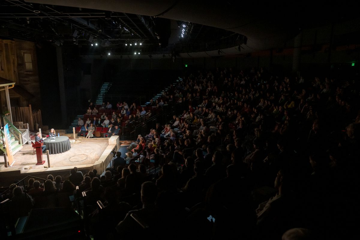 Stage with two people speaking, audience seated in a darkened theater, some lights on stage, audience in shadows.