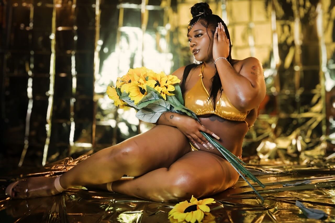 Woman in gold swimsuit sitting in water holding a sunflower bouquet, surrounded by bamboo and sunlight.