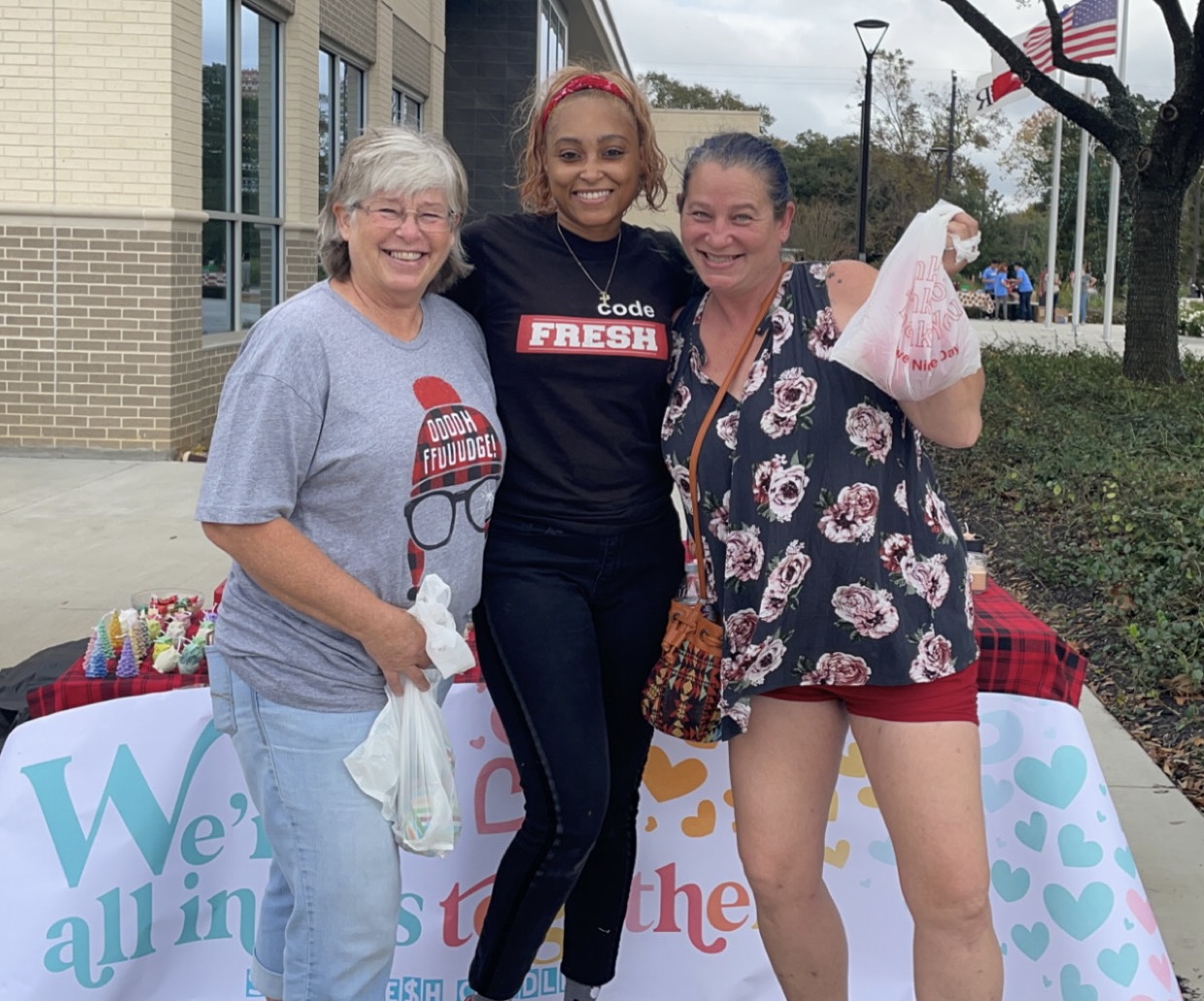 Three women smiling outdoors, one holding a pink stuffed animal, standing near a table with a colorful banner.