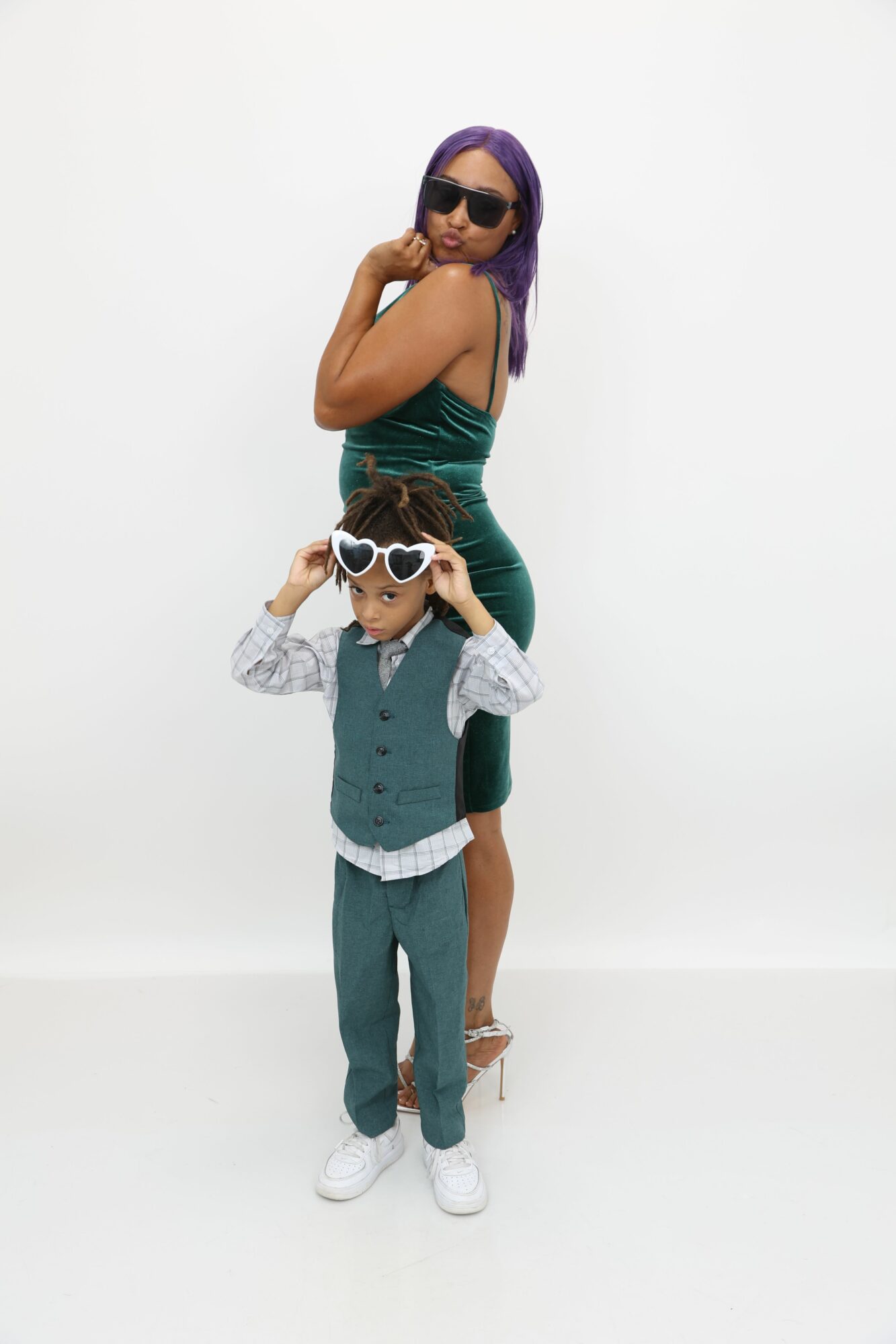 Person with purple hair and sunglasses taking a selfie with a woman in a green dress, against a plain white background.