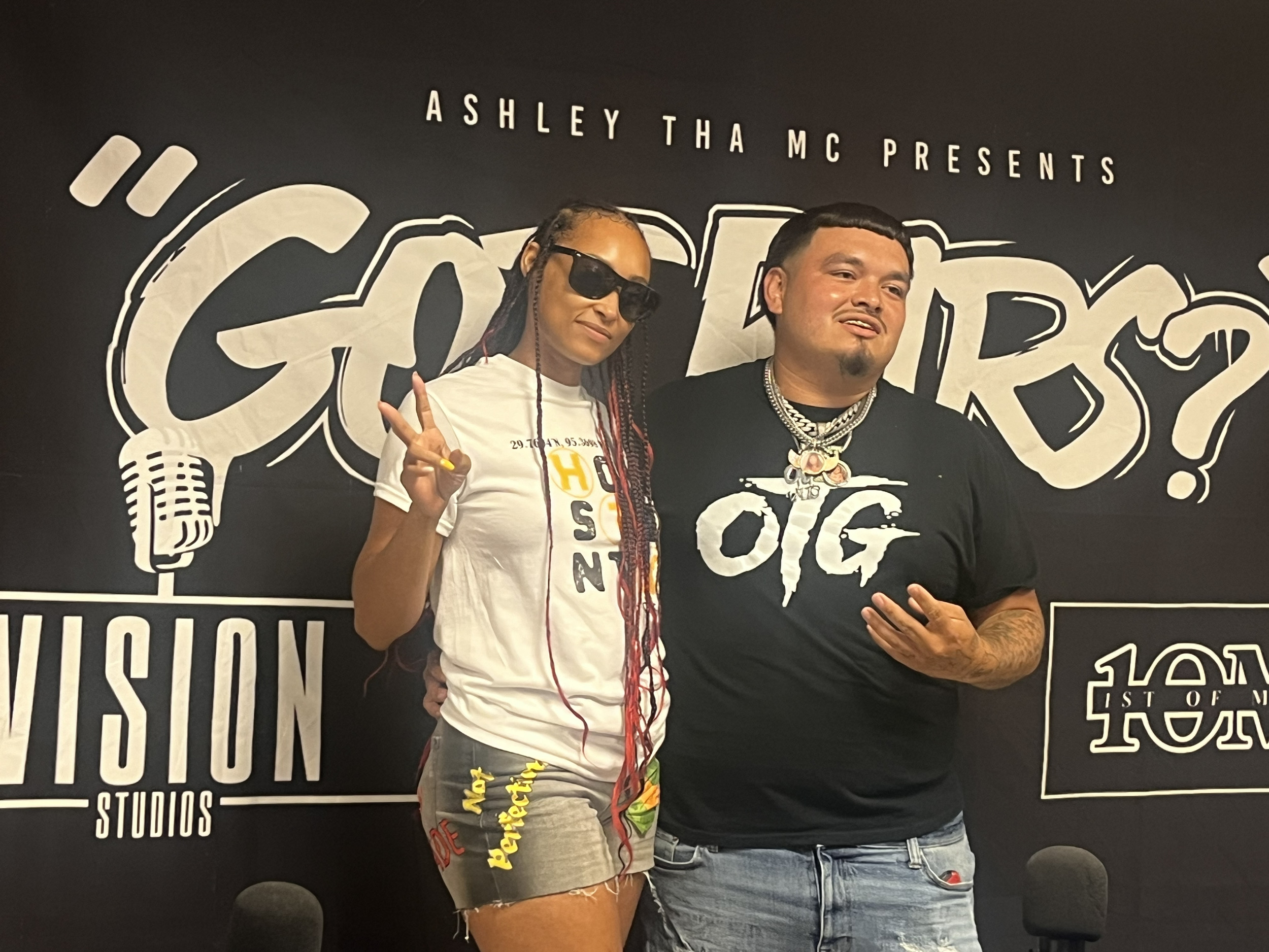 Two people standing in front of a black background with text, one wearing sunglasses and a white shirt, the other in a black shirt.