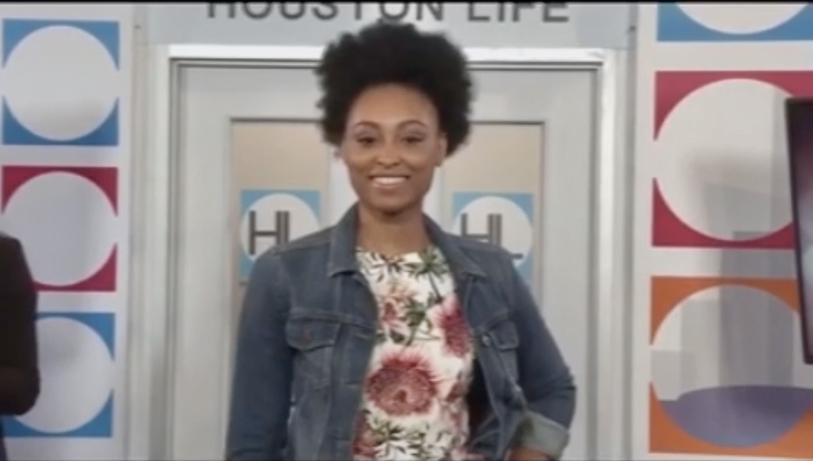 Smiling woman with short curly hair wearing a denim jacket and floral top, standing in front of a colorful background.