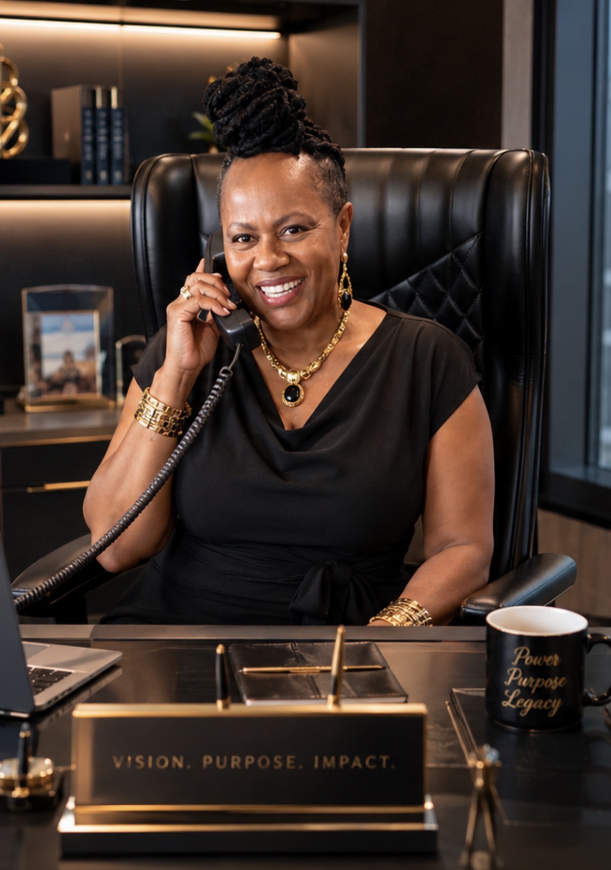 Woman sitting at desk, smiling, talking on phone, in a modern office with books and a mug nearby.
