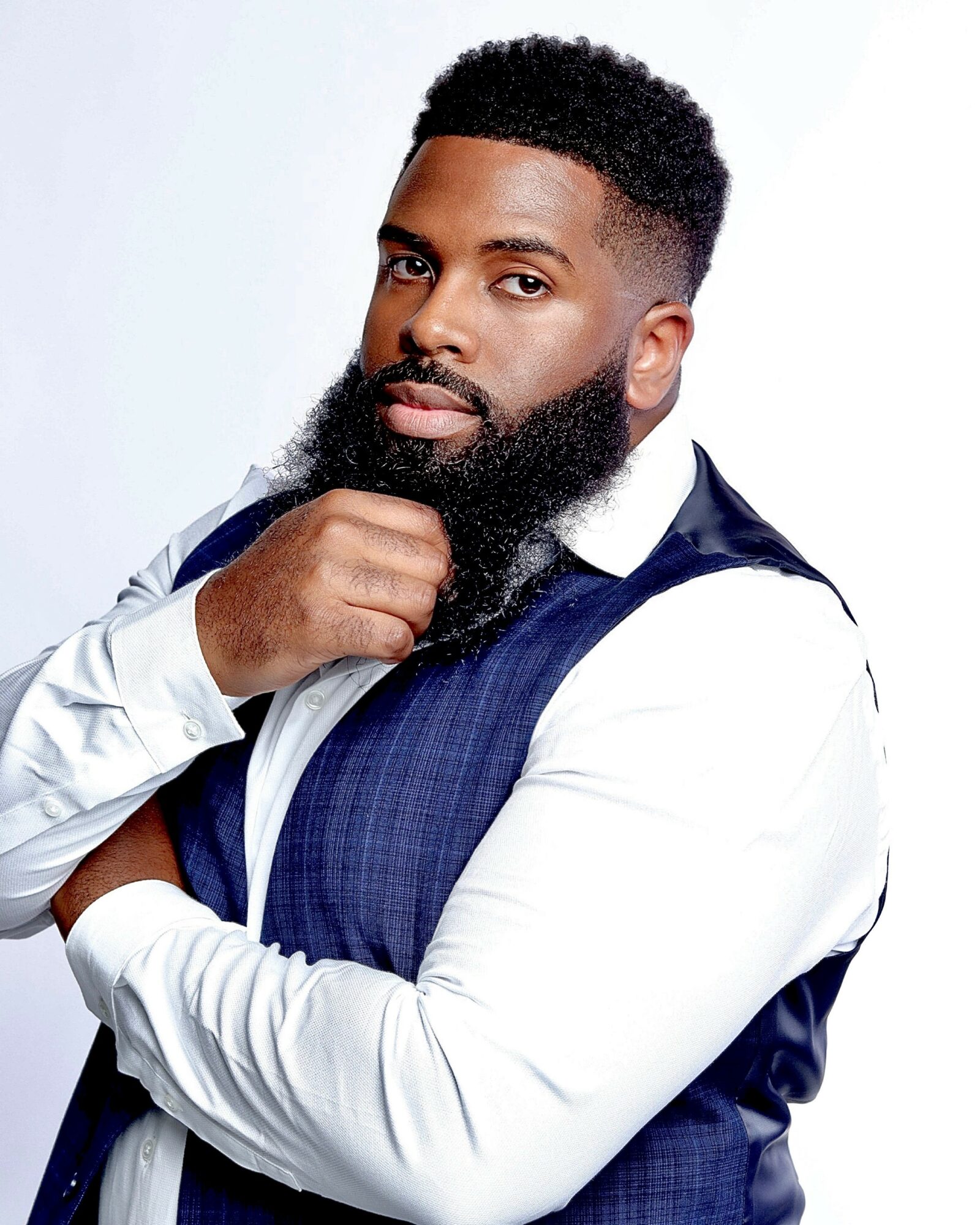 Man with a beard and short curly hair, wearing a white shirt and blue vest, posing with hand on chin.