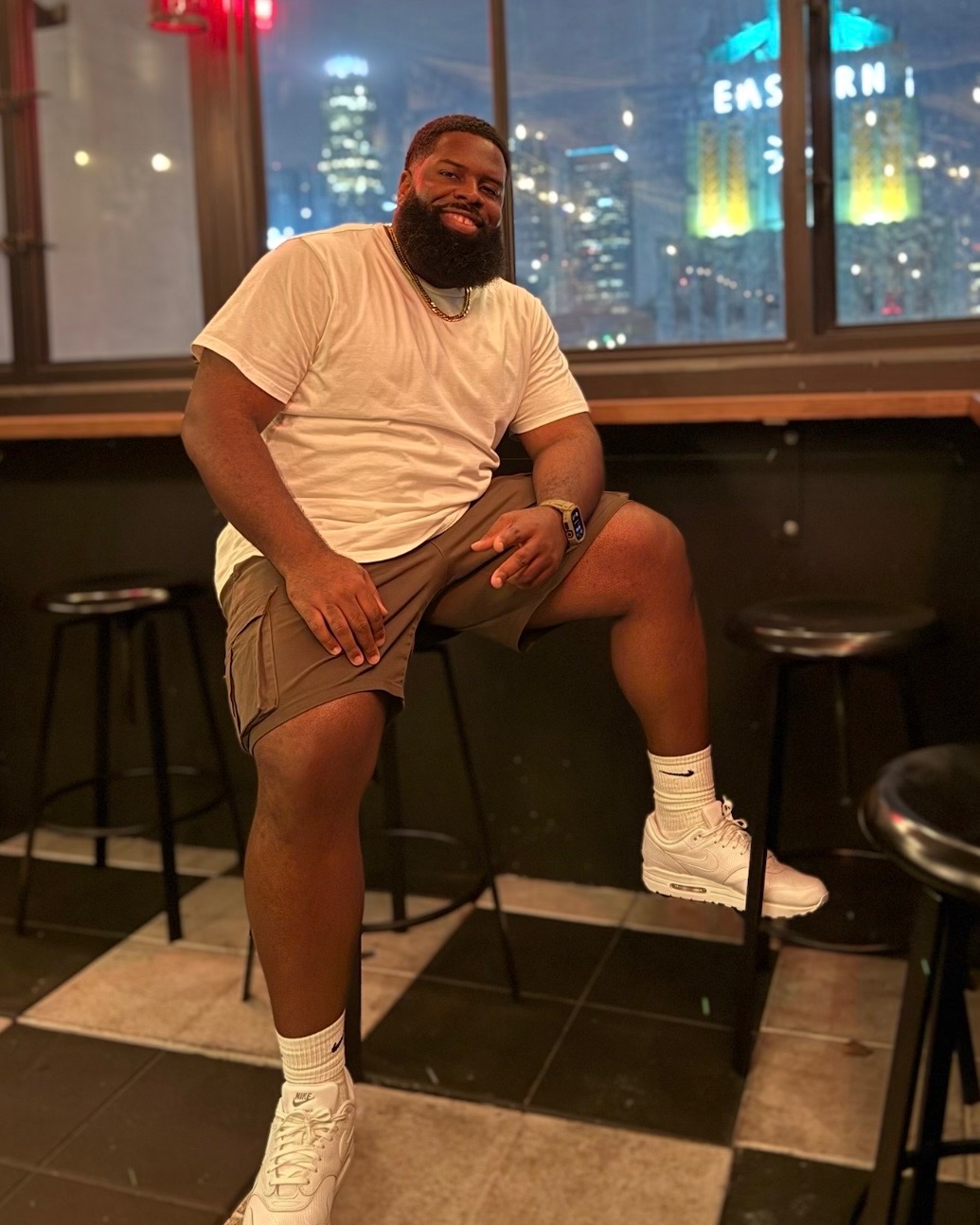 Man with a beard smiling, sitting on a high stool in a room with large windows and city lights outside.