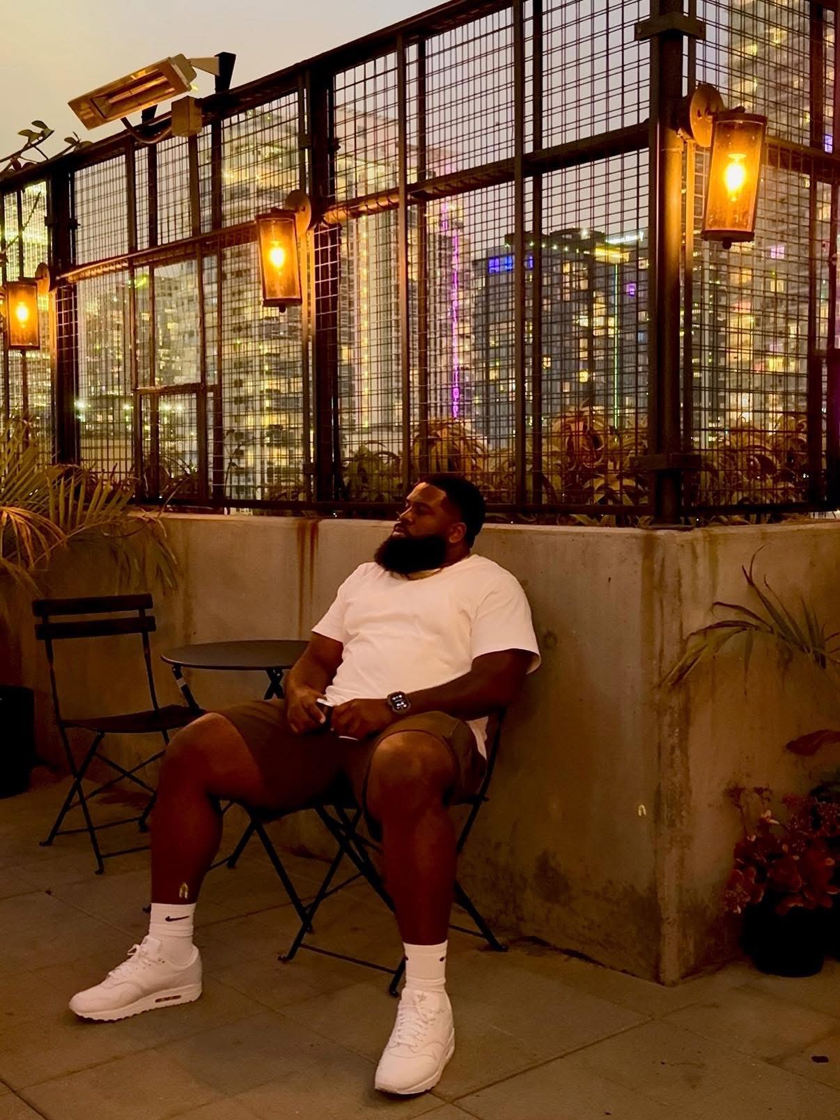 Man sitting on a chair outdoors at night with city lights and warm lighting in the background.