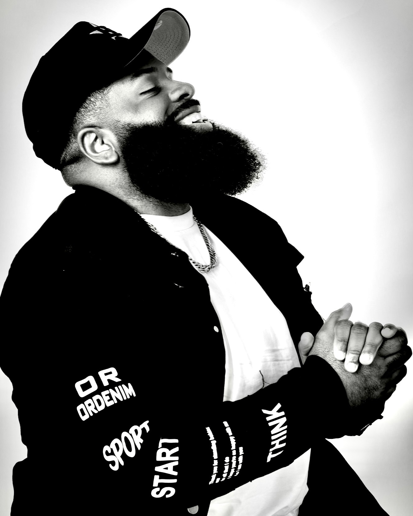 Man with a beard and cap smiling, wearing a jacket with text, clasping hands, against a plain background.