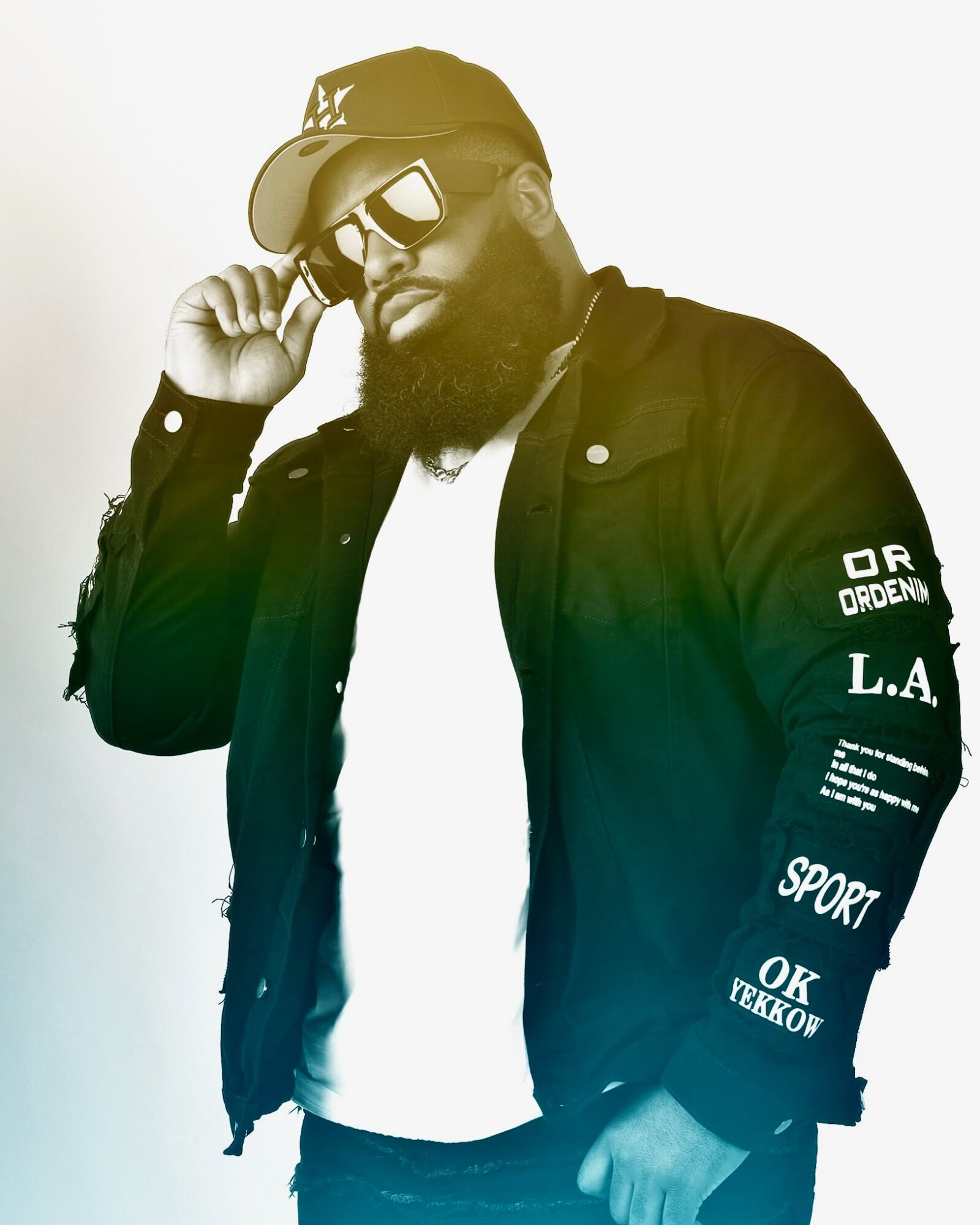 Man with beard wearing sunglasses, cap, and jacket, adjusting his sunglasses, standing against a plain background.