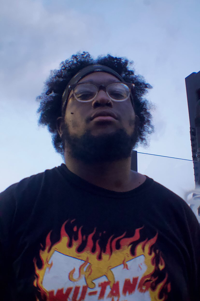 Young person with glasses and curly hair outdoors, wearing a black T-shirt with a flaming graphic, against a cloudy sky.