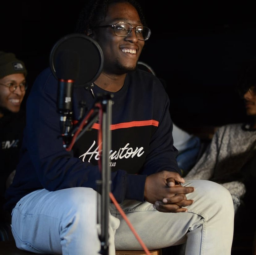 Person smiling, sitting with hands clasped, in a recording studio with a microphone, wearing glasses and a dark sweatshirt.