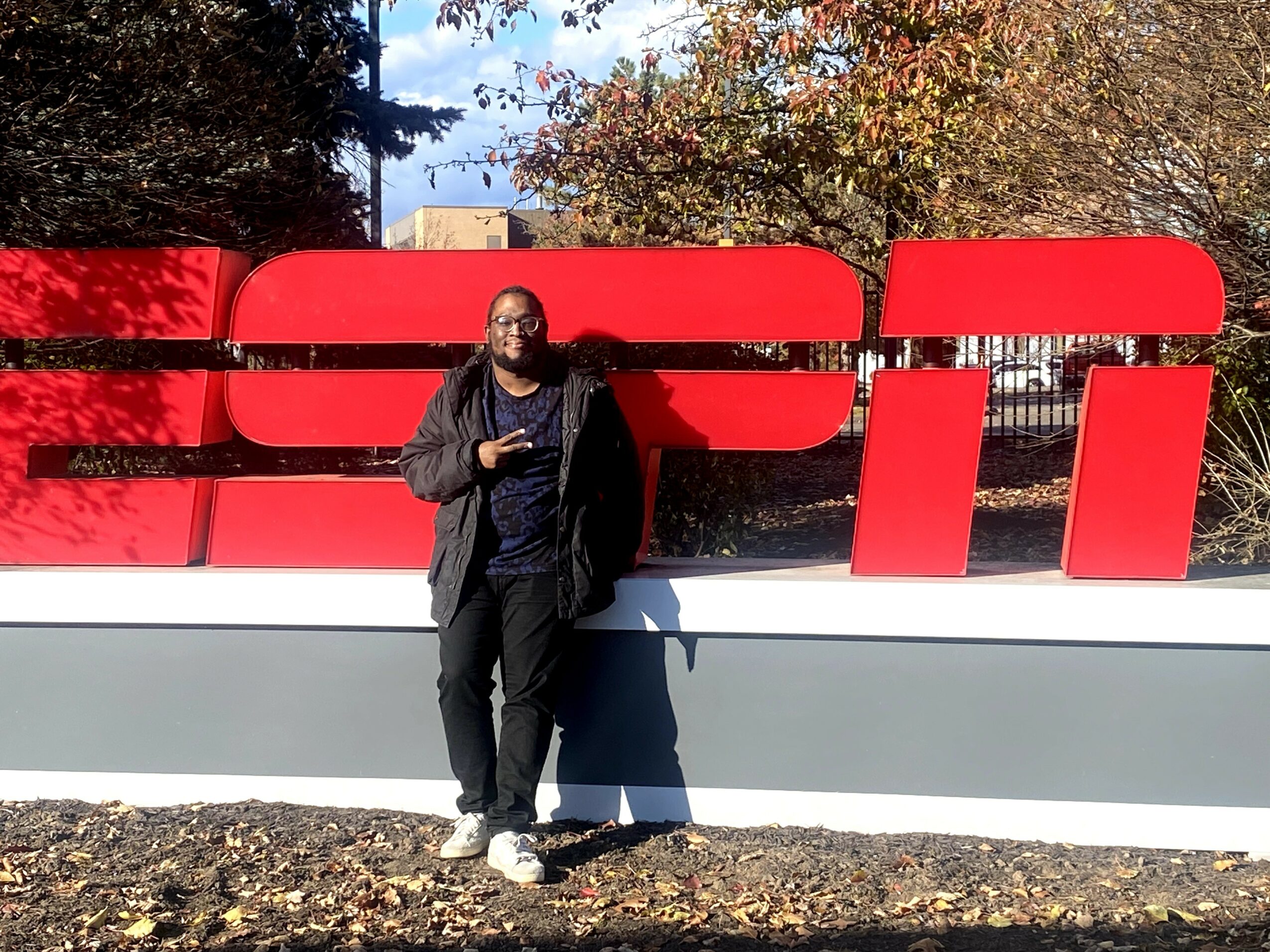 Person standing in front of a large red and white sign outdoors, with trees and sky in background.