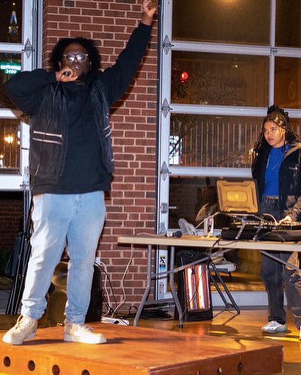 Person with glasses and black jacket singing into a microphone, raising one arm, in front of a brick wall and glass window.