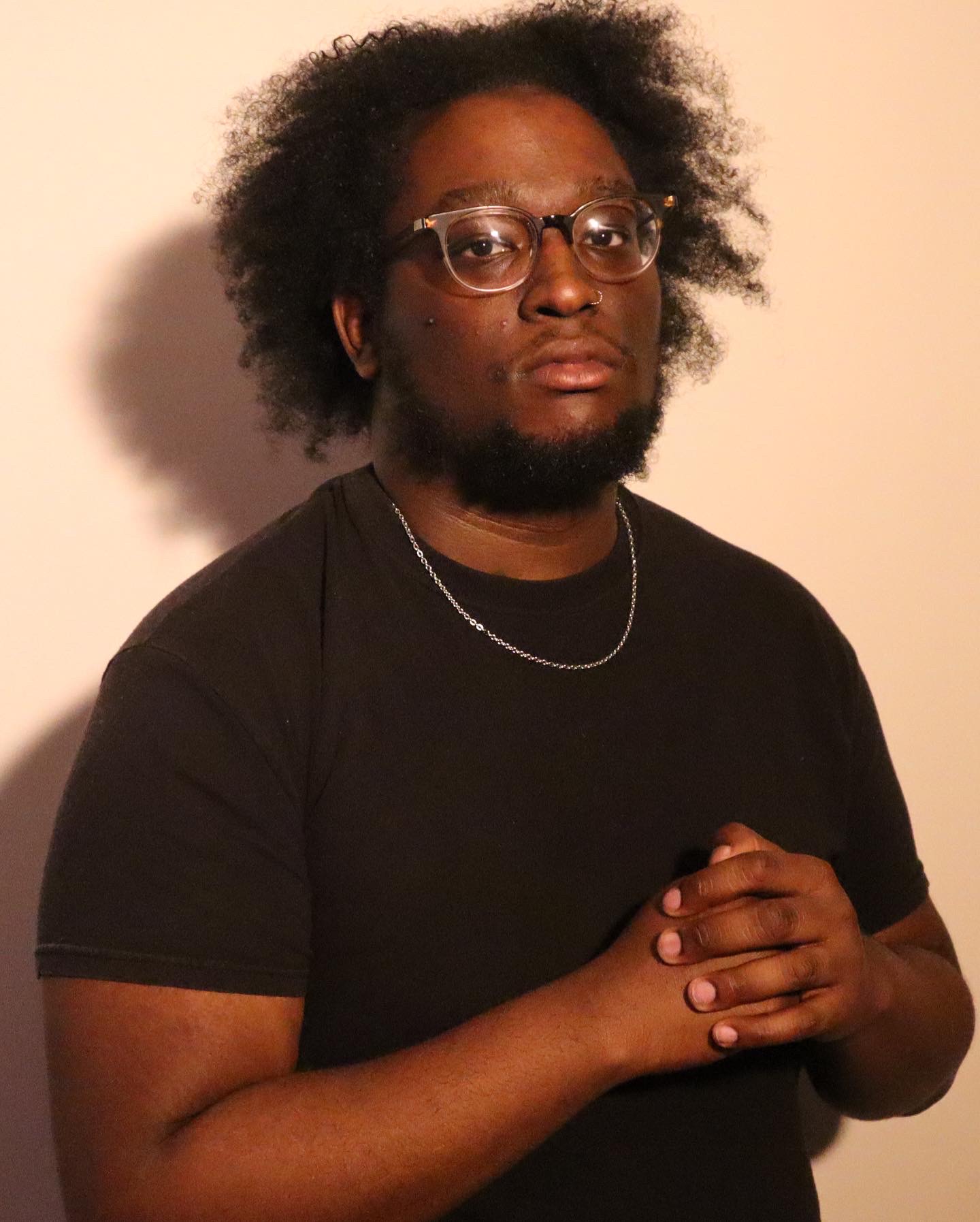 Person with glasses and curly hair, wearing a black shirt, standing against a plain background.