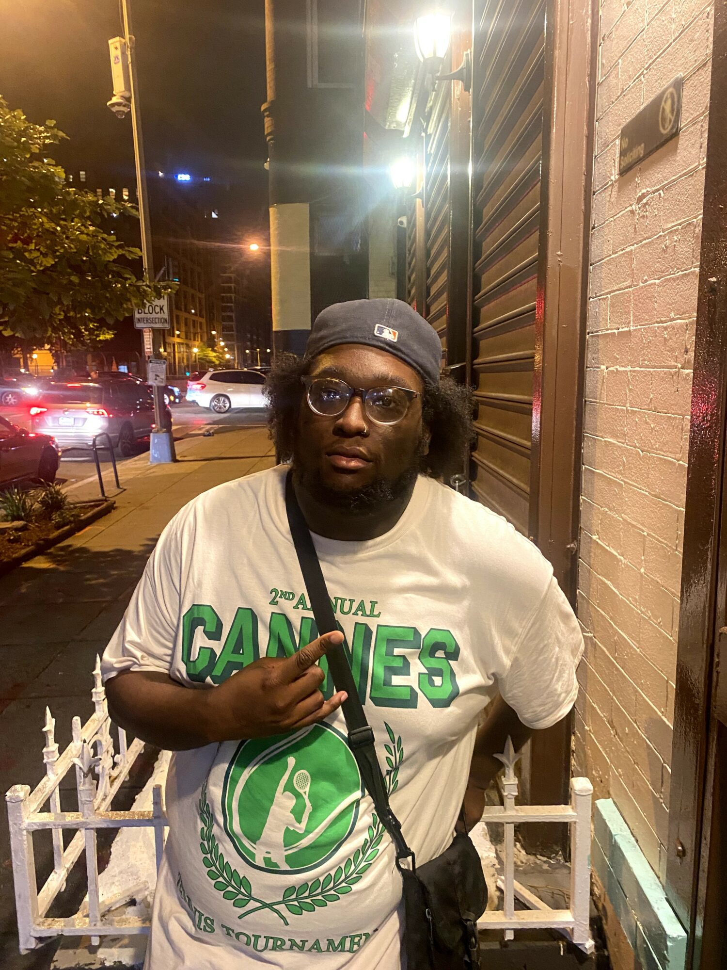 Young man with glasses and a cap standing outside at night, wearing a white T-shirt with green text and graphics.