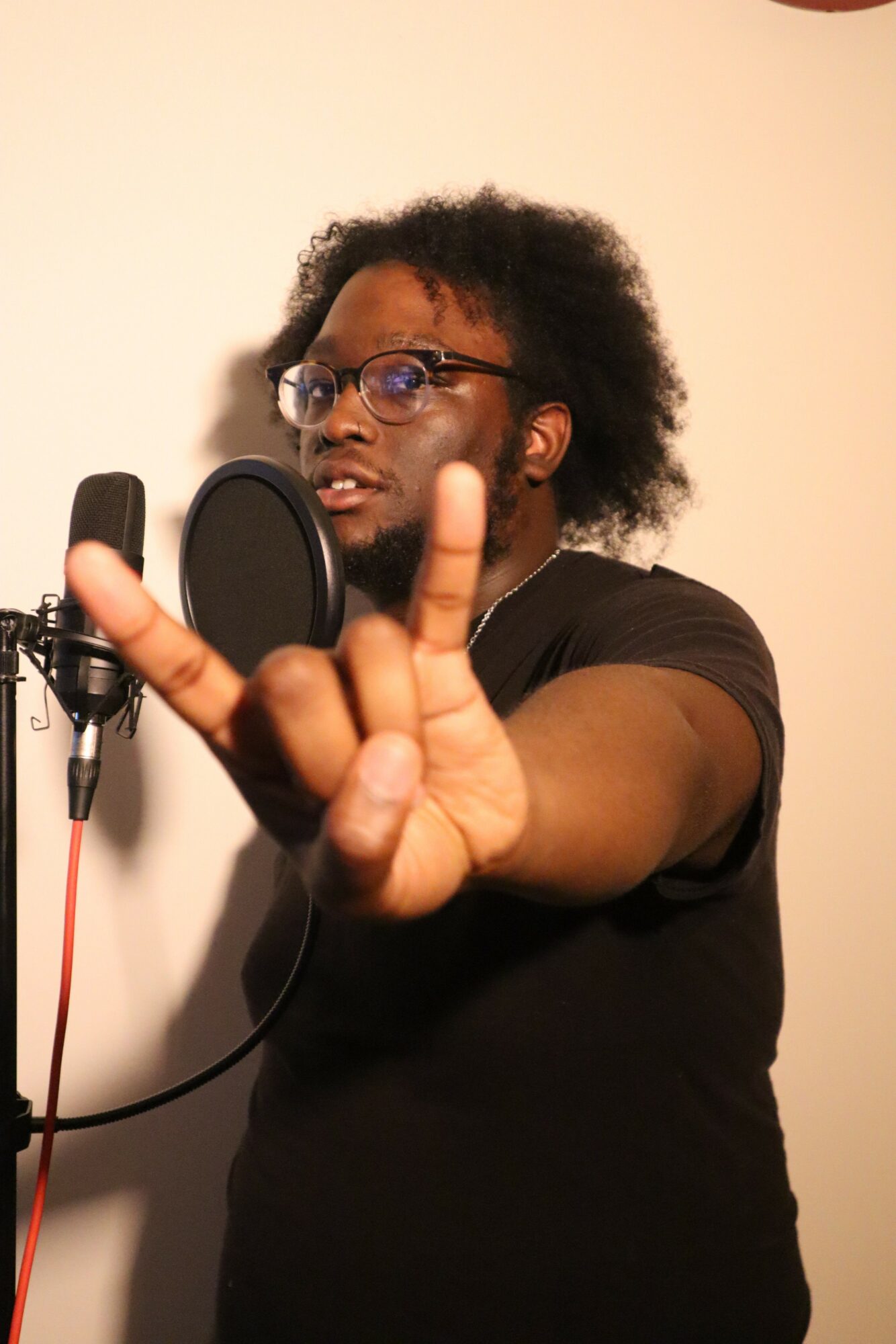 Person with glasses and curly hair recording into a microphone, making a hand gesture, against a plain background.