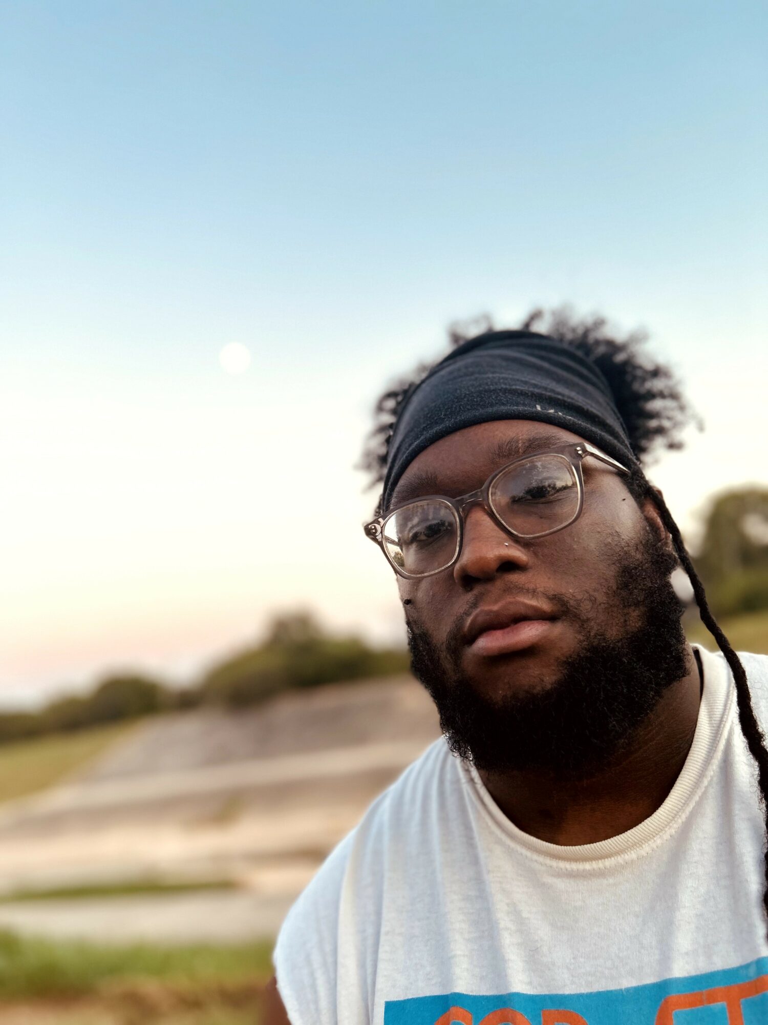 Young man with glasses and headband outdoors, sky and landscape in background.