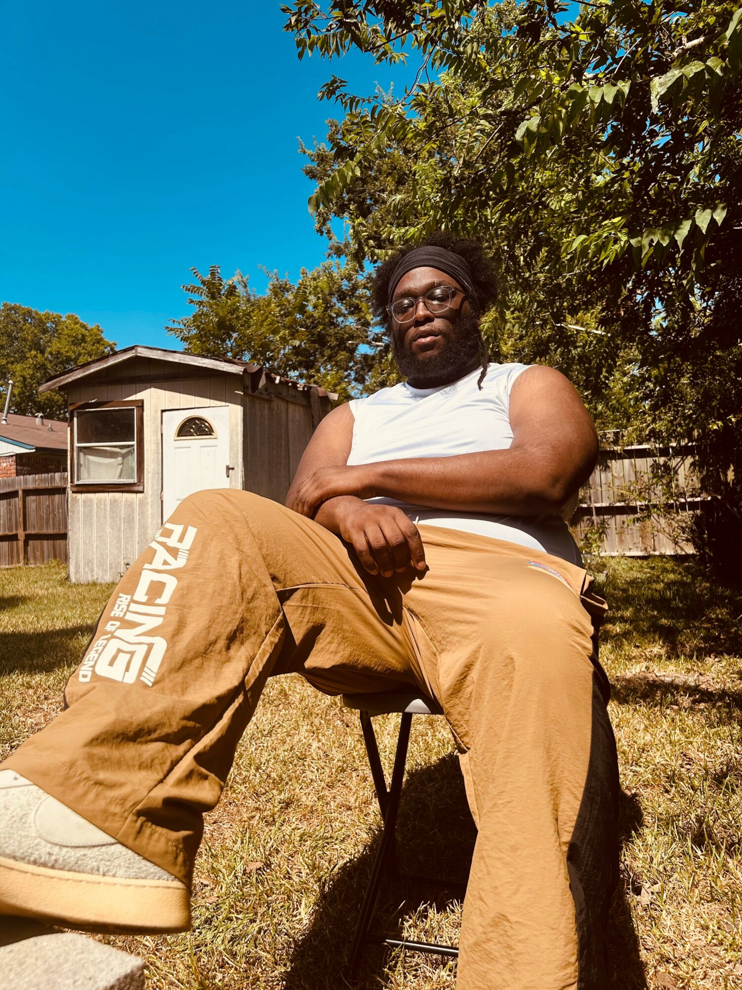 Person sitting outdoors on a chair, wearing glasses, a white sleeveless top, and tan pants with white text, in a yard with trees and a wooden fence.