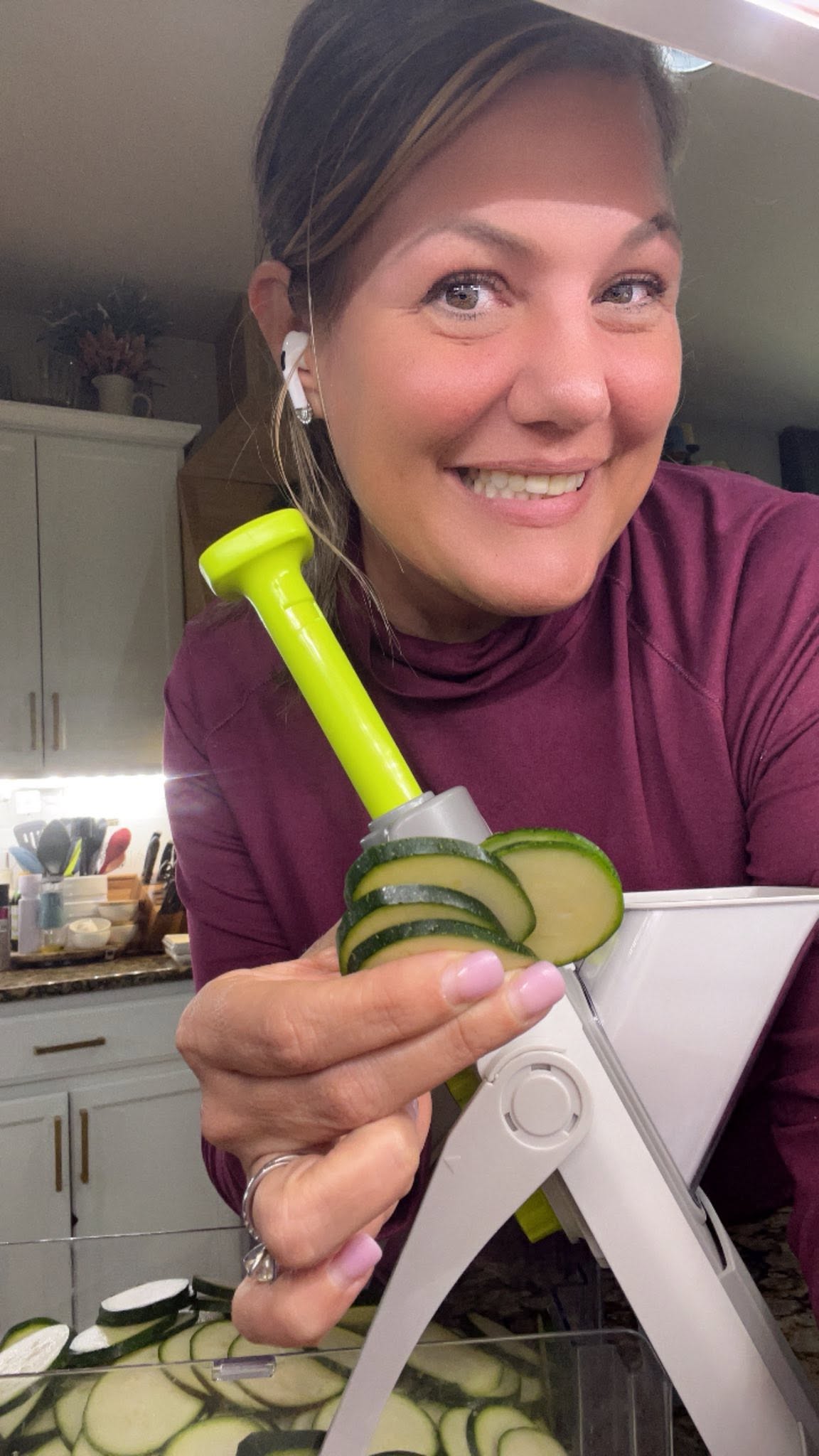 Woman smiling in kitchen holding a vegetable slicer with cucumber slices, wearing earphones and a maroon top.