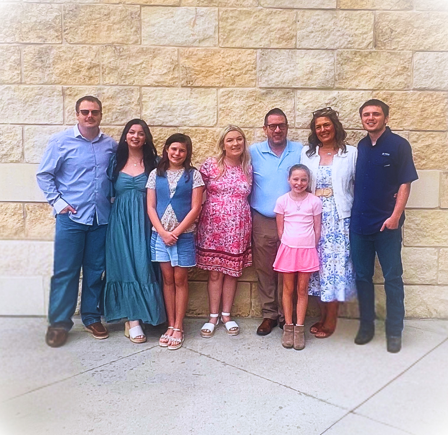 Group of eight people standing against a stone wall, smiling, with a mix of adults and children, dressed casually.