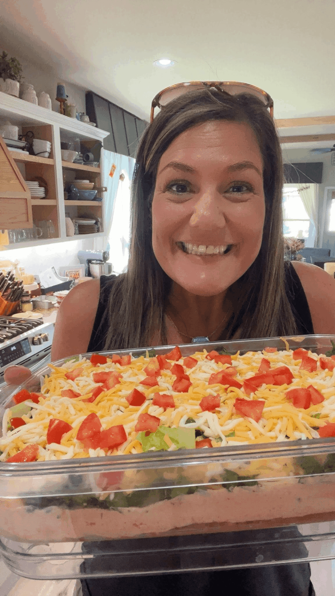 Smiling woman with long brown hair in a kitchen, holding a dish with colorful toppings, including red and yellow pieces.