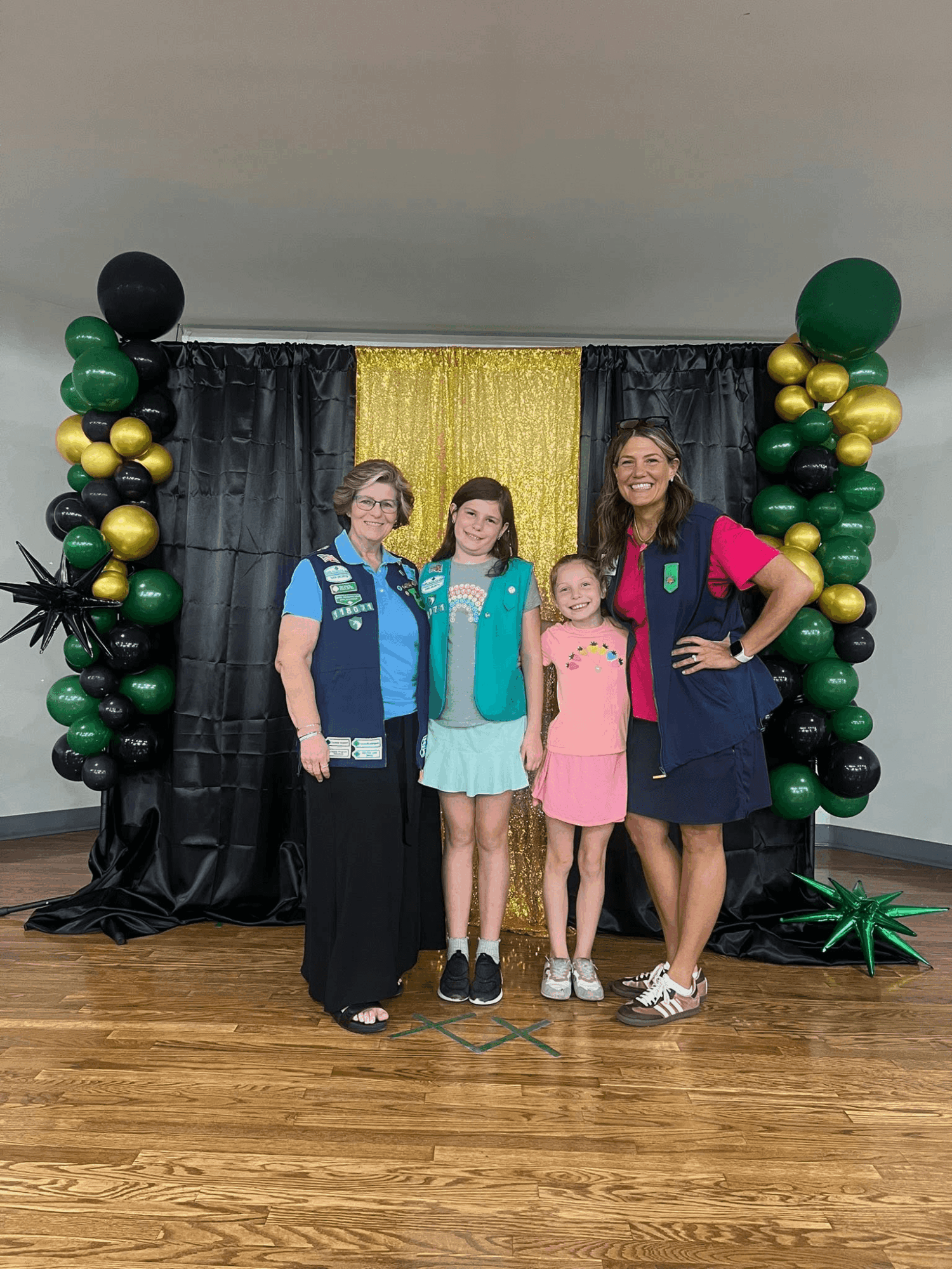 Four people stand in front of a decorated backdrop with black, gold, and green balloons and a gold curtain, smiling.