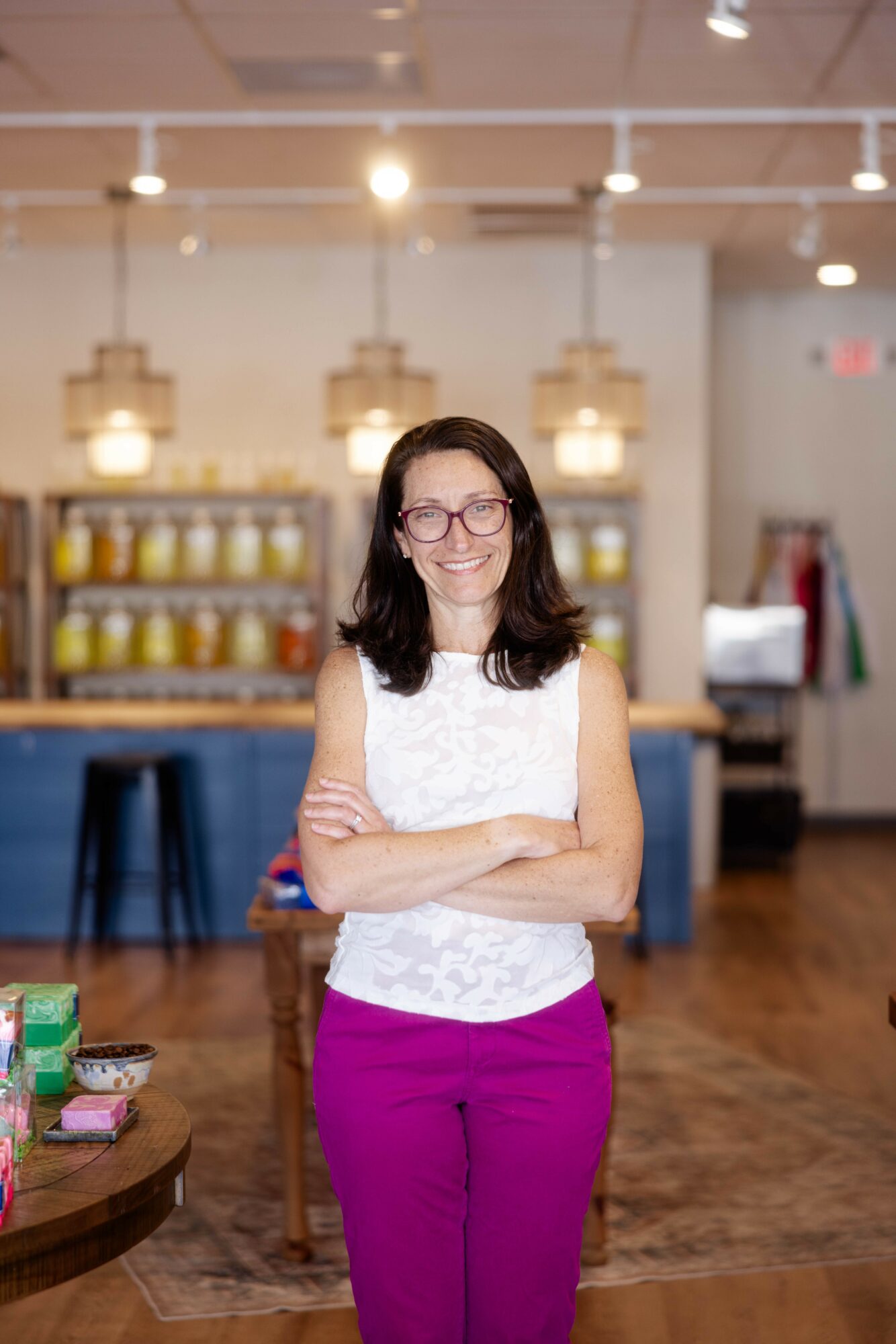 Woman with glasses and dark hair standing with arms crossed in a store or cafe, background shelves and lighting fixtures.