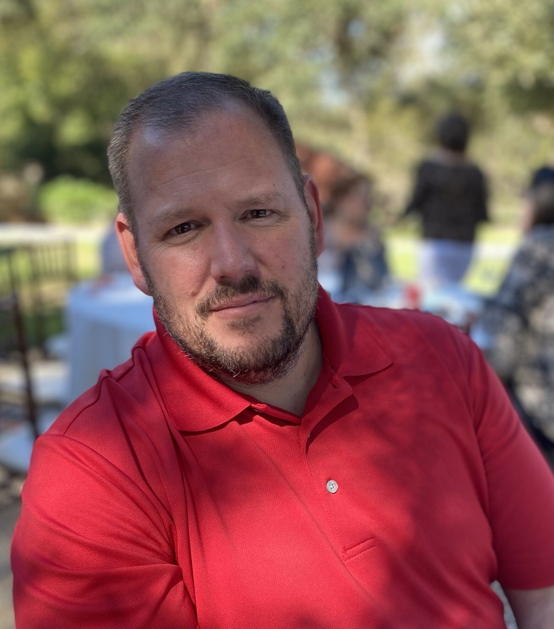 Man with short hair and beard wearing a red polo shirt outdoors, smiling, with trees and people in the background.