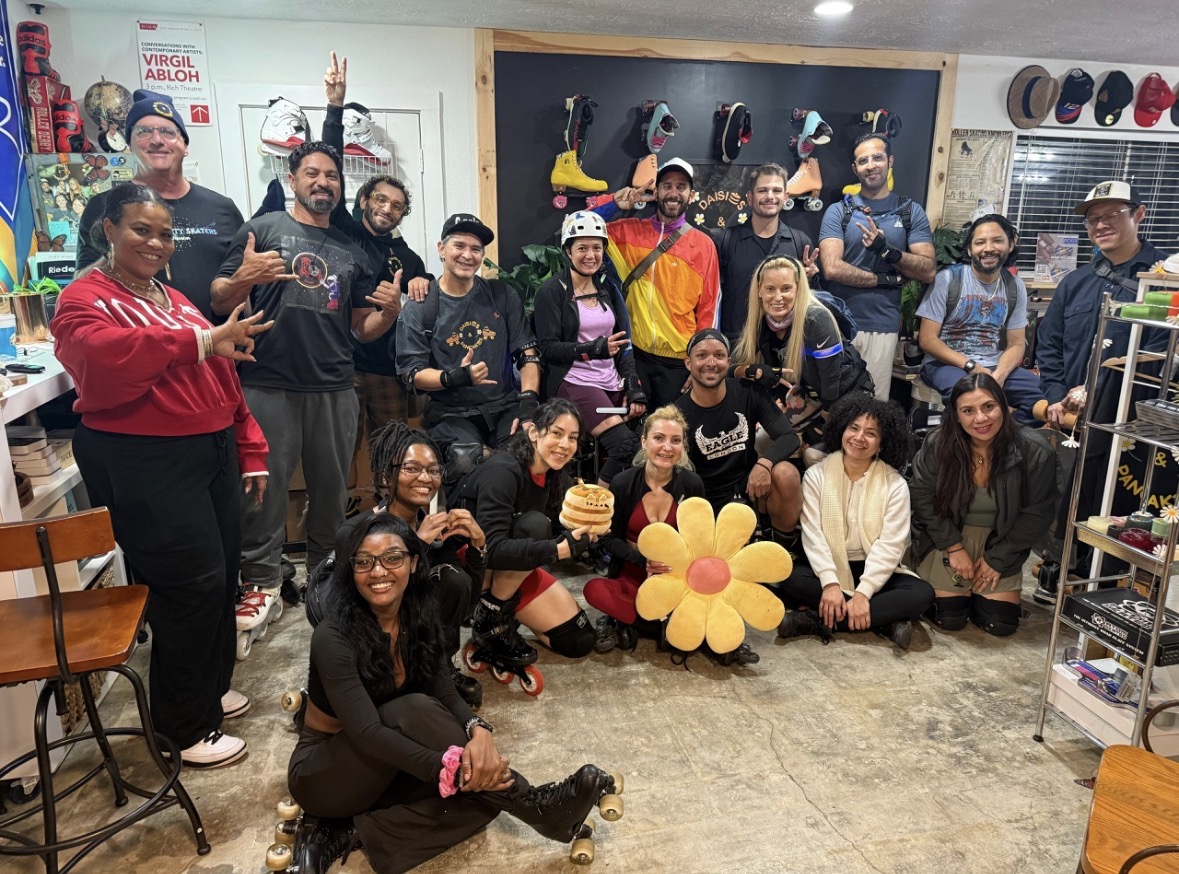 Group of people indoors, some wearing helmets, holding a large flower prop, smiling, with climbing gear and helmets on the wall behind.