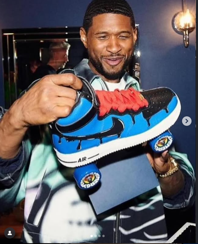 Man holding a blue and black sneaker with red laces, smiling, in a room with dark walls and a light fixture.