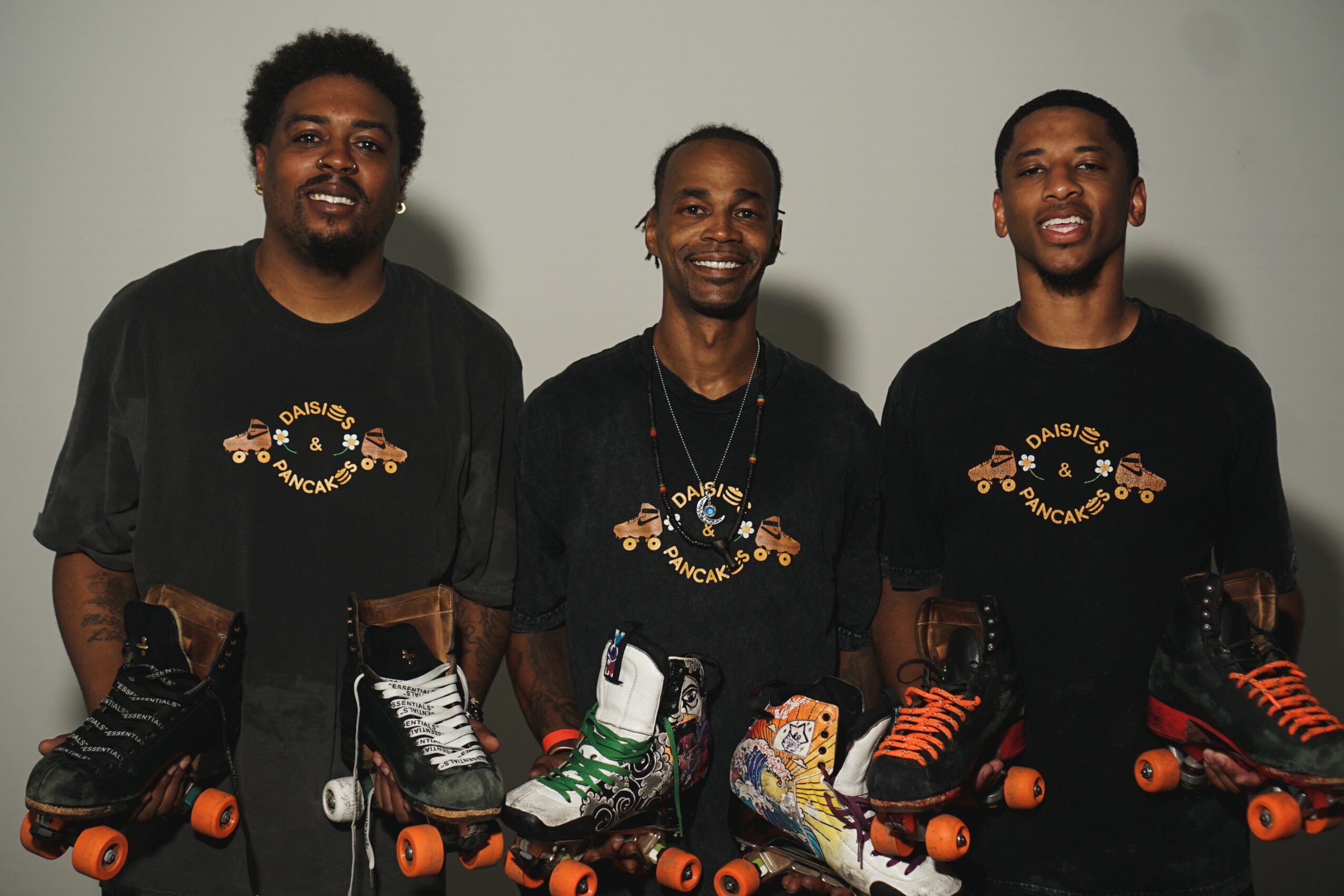 Three people standing together, each holding roller skates, smiling, wearing black shirts with a circular logo, against a plain background.