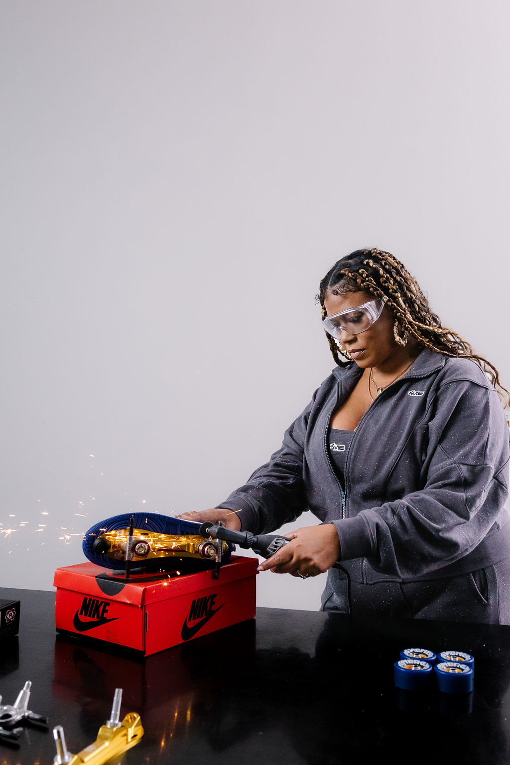 Woman wearing safety glasses and a jacket, holding a blowtorch over a red Nike shoe box, with small blue objects nearby.