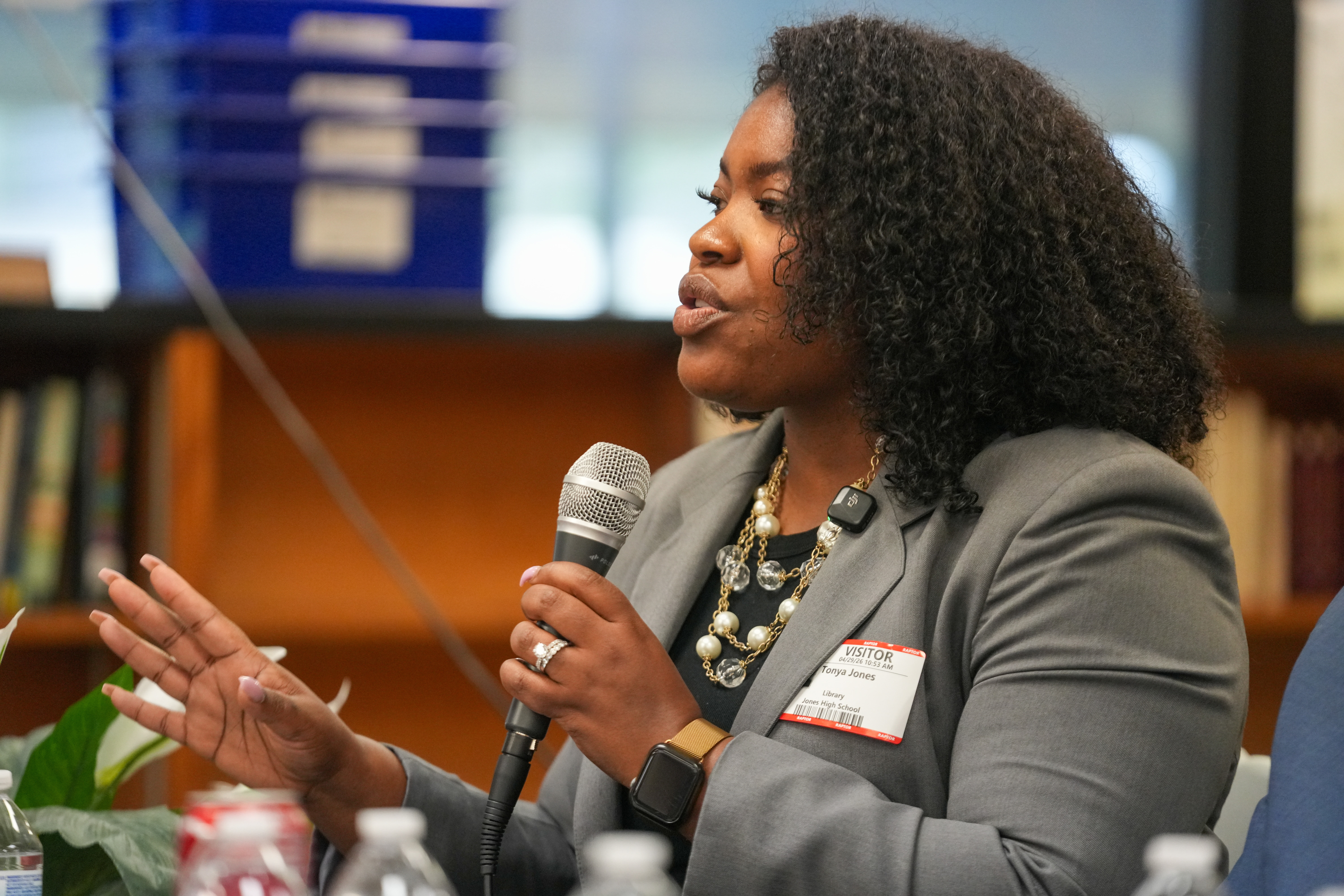 Woman speaking into a microphone, gesturing with her hand, in a professional setting with bookshelves in the background.