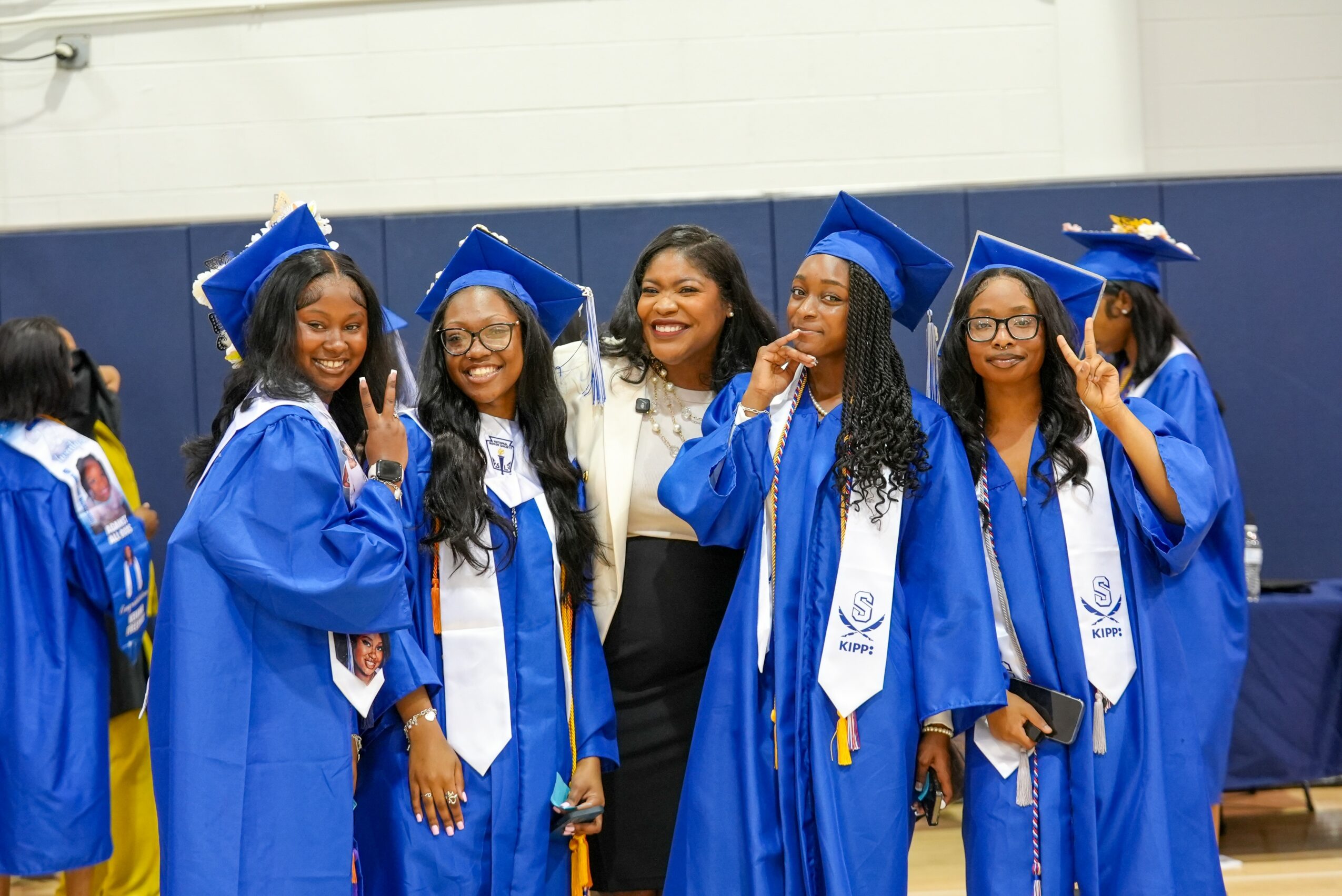 Group of five young women in blue graduation gowns and caps, smiling and posing indoors with a woman in white blazer, celebrating graduation.
