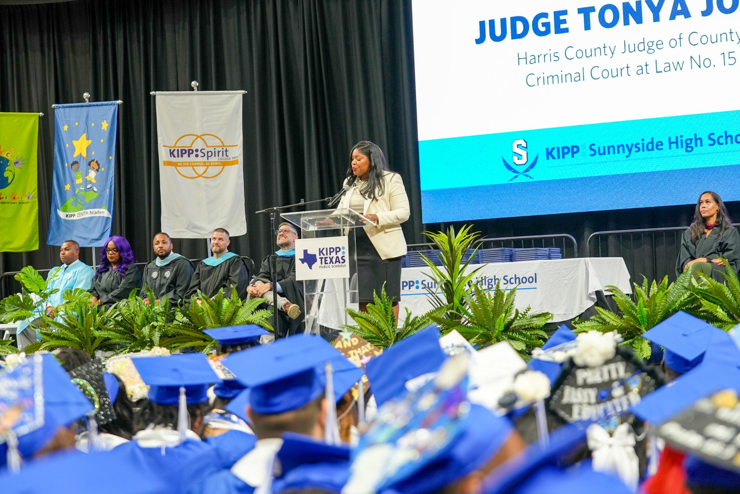Woman speaking at podium during graduation ceremony with seated graduates in caps and gowns, stage with banners and large screen in background.