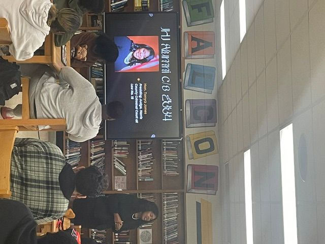 People seated in a room with a large screen displaying a presentation and bookshelves in the background.