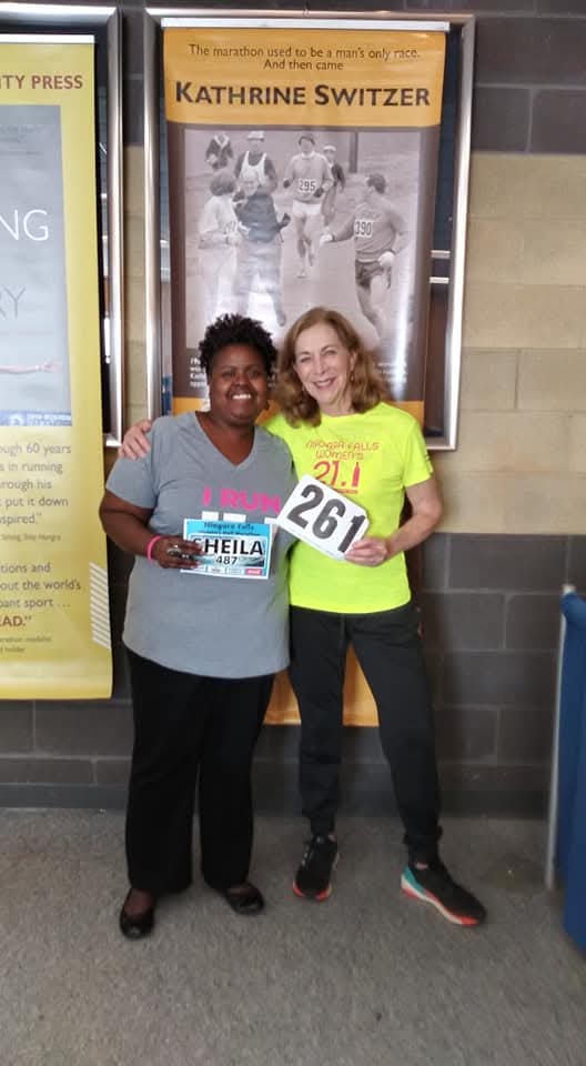 Two women standing together, smiling, one holding a race bib with the number 267, in front of a poster and brick wall.