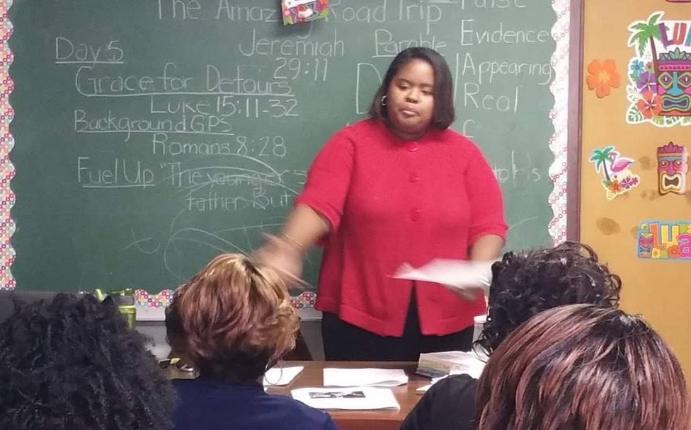 Teacher standing in front of a classroom blackboard, speaking to students seated at desks, holding papers, classroom decorations visible.