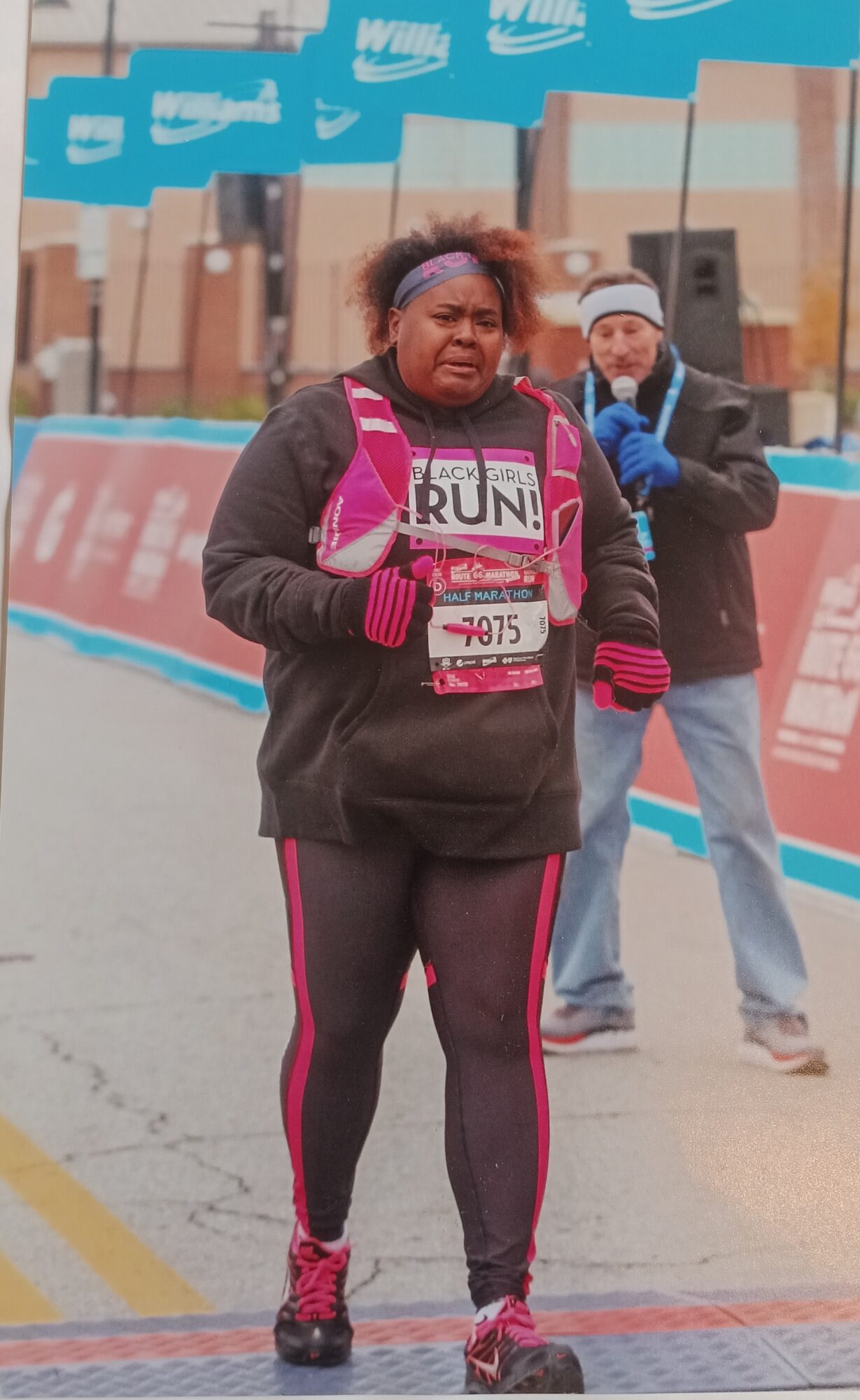 Woman in black and pink athletic clothing participating in a race, with a man in the background, outdoors.