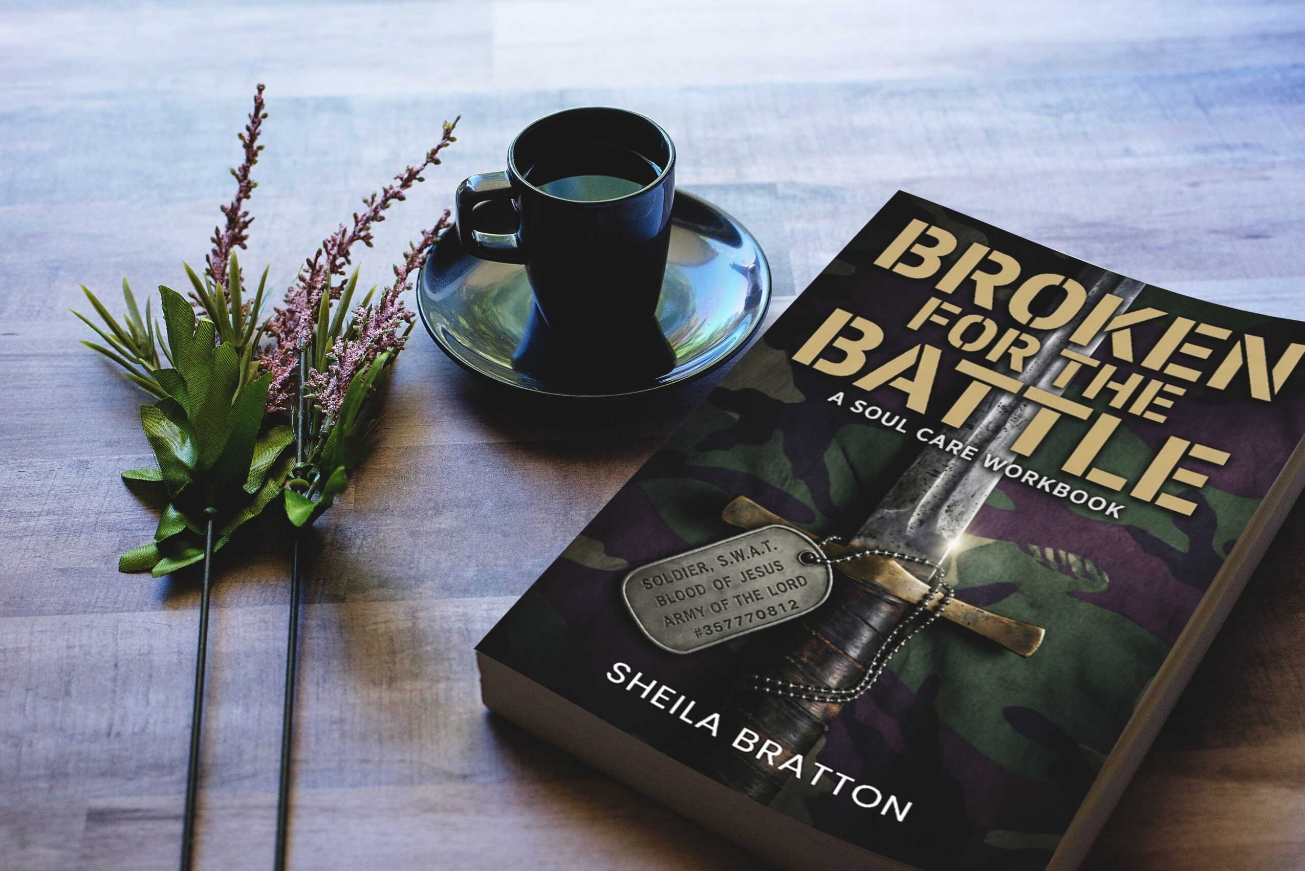 A book titled 'Broken for the Battlefield' by Sheila Bratton, a black coffee cup on a saucer, and a small bouquet of flowers on a wooden surface.
