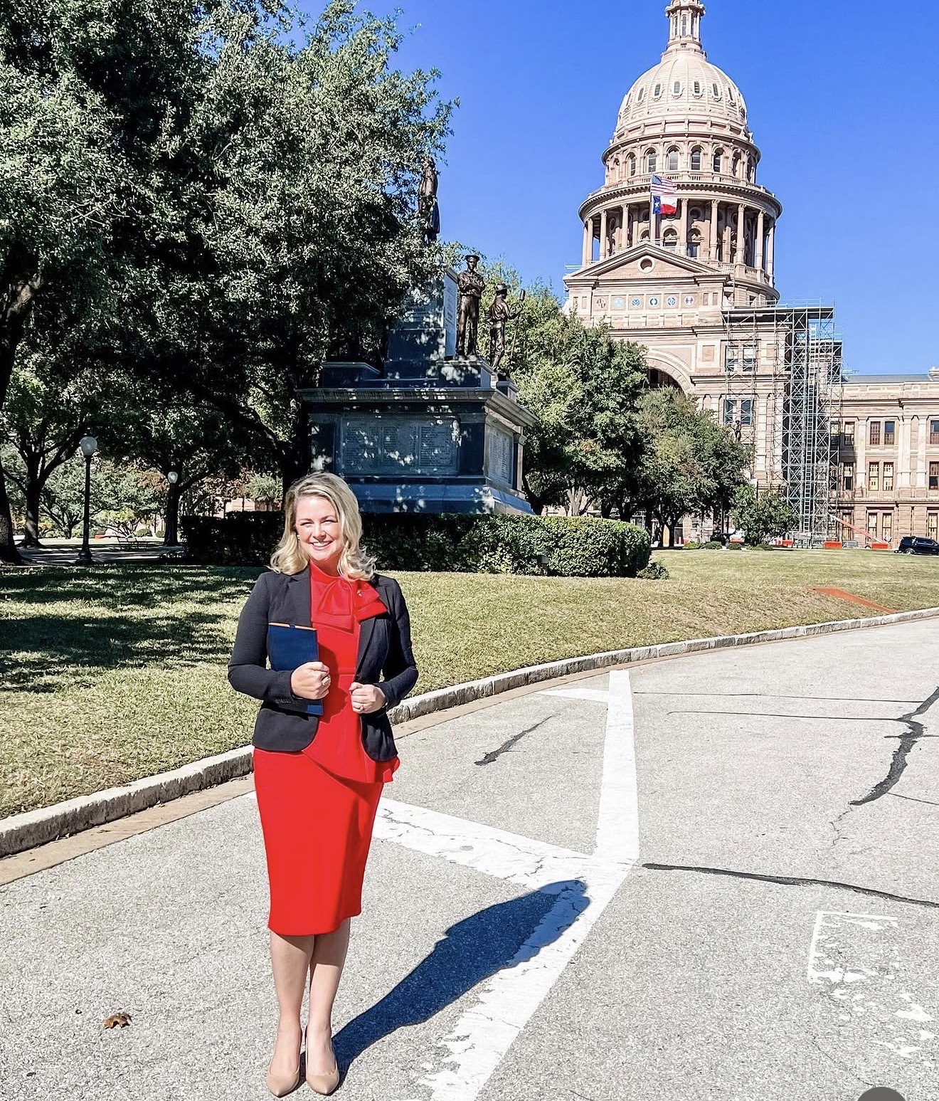 Woman in red dress and black blazer standing outdoors near a historic building with a dome, trees, and a monument, sunny day.