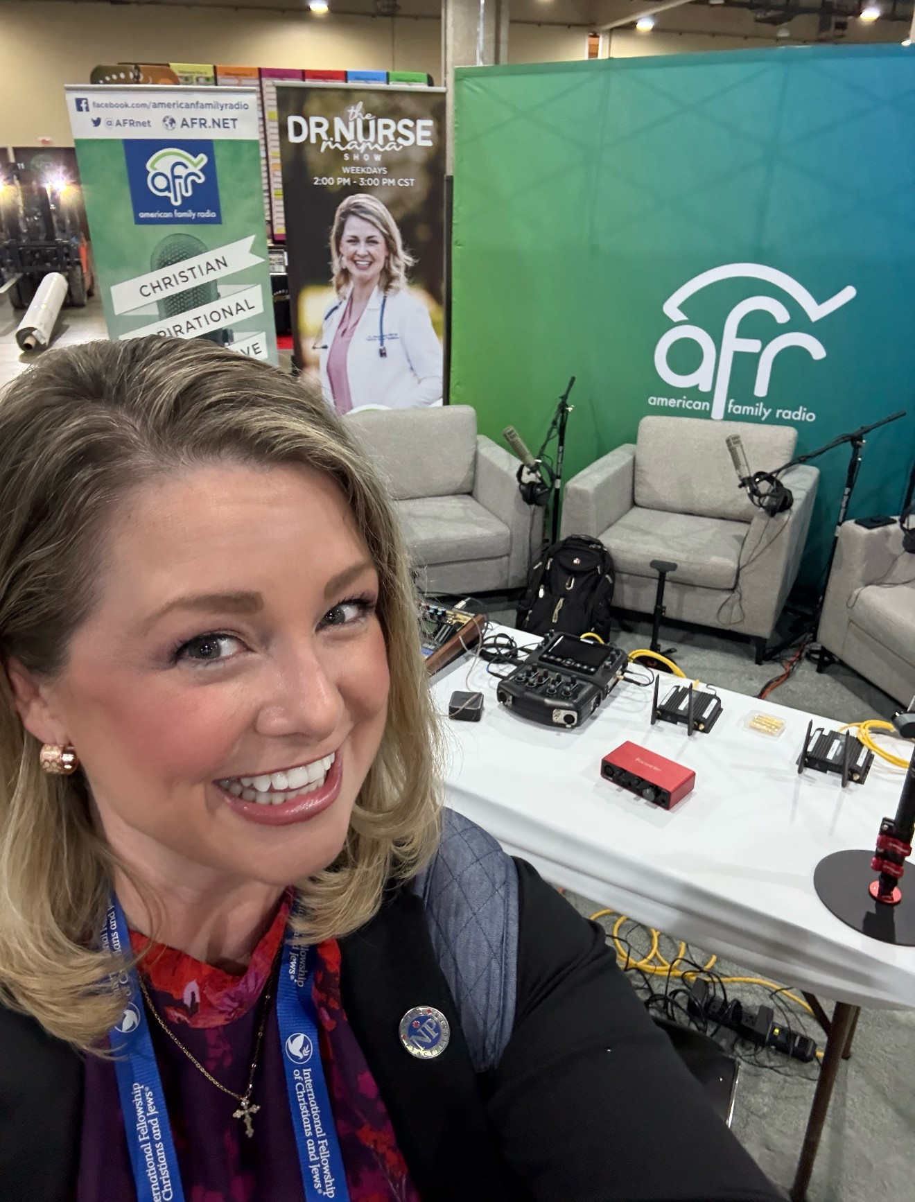Smiling woman with blonde hair taking selfie in radio studio with microphones and chairs, green and blue background with logo.