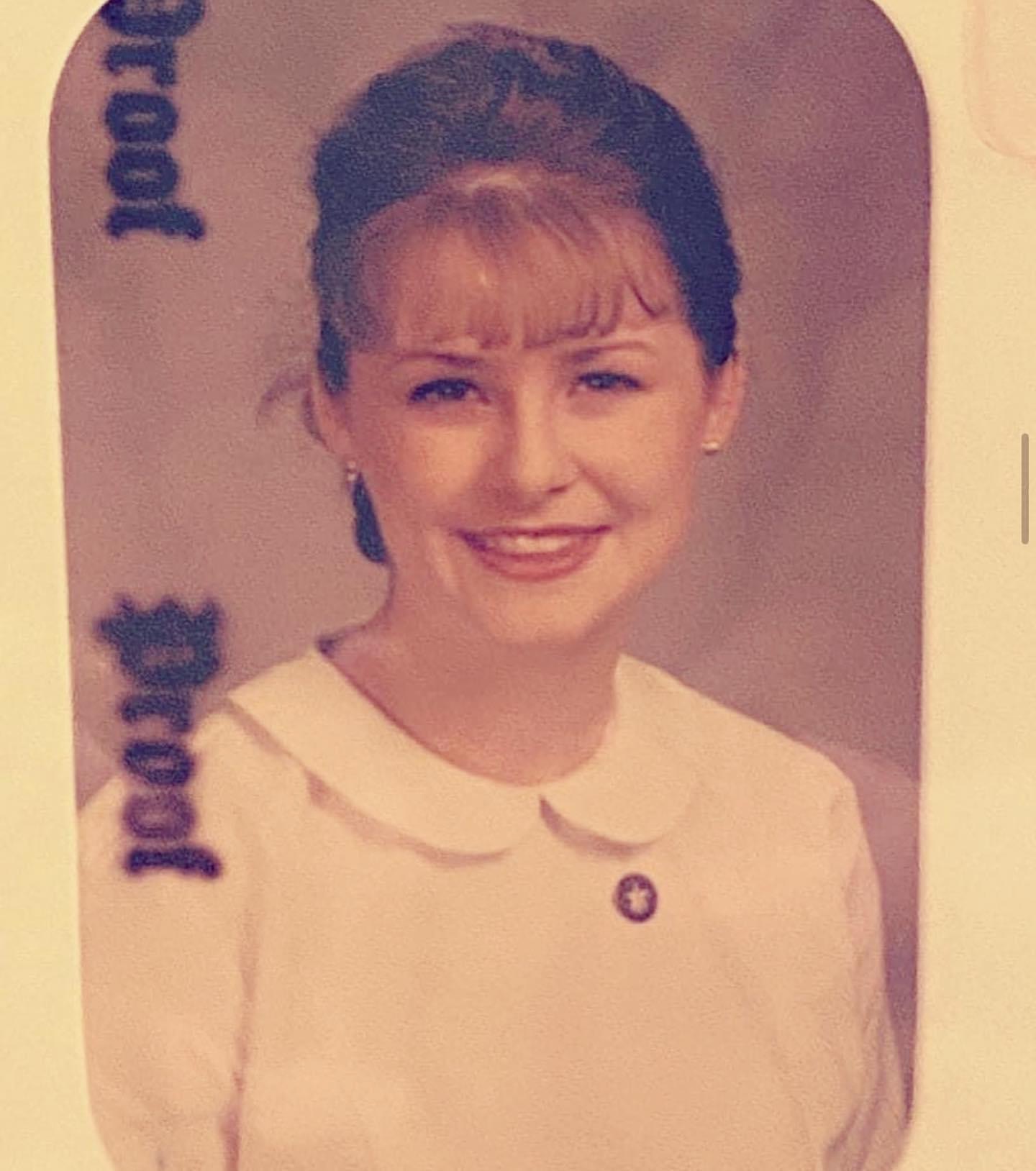 Young woman with light brown hair styled in an updo, wearing a white collared dress, smiling, with a blurred background.