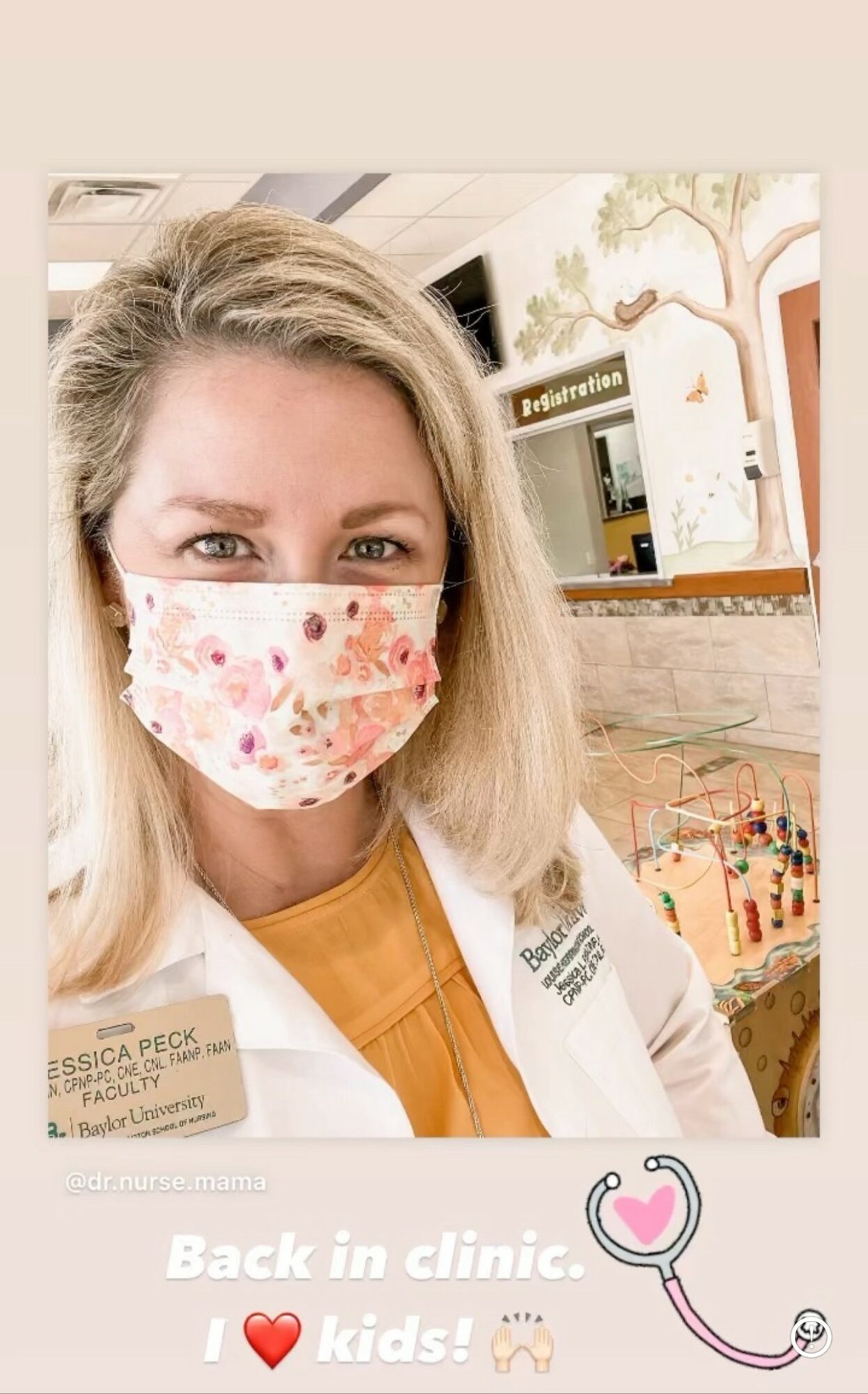A woman wearing a floral face mask and white coat in a clinic, with a colorful play area in the background.