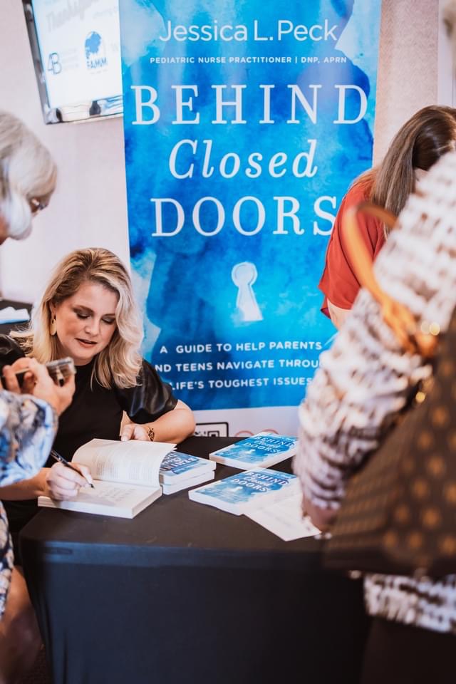 Author Jessica L. Peck signing books at a table with a blue book cover behind her, surrounded by people.