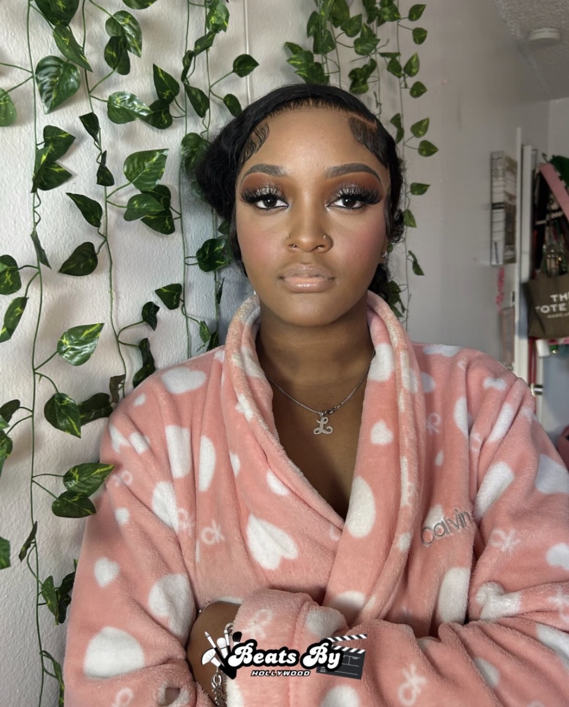 Young woman with makeup and styled hair, wearing a pink and white polka dot robe, standing in front of green leafy plants.